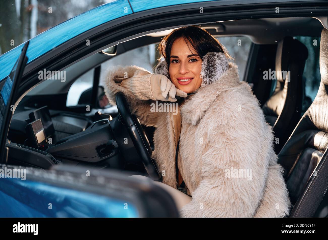 La donna con i capelli lunghi, vestita con un elegante cappotto di pelliccia, è seduta in un'auto, trasuda calore e gioia in un ambiente invernale con illuminazione soffusa Foto Stock