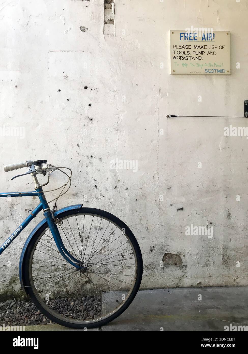 Una bicicletta Triumph d'epoca si appoggia contro un muro battuto dal tempo e presenta una stazione pubblica "Free Air" e una stazione di attrezzi per biciclette in Scozia, Regno Unito. Foto Stock
