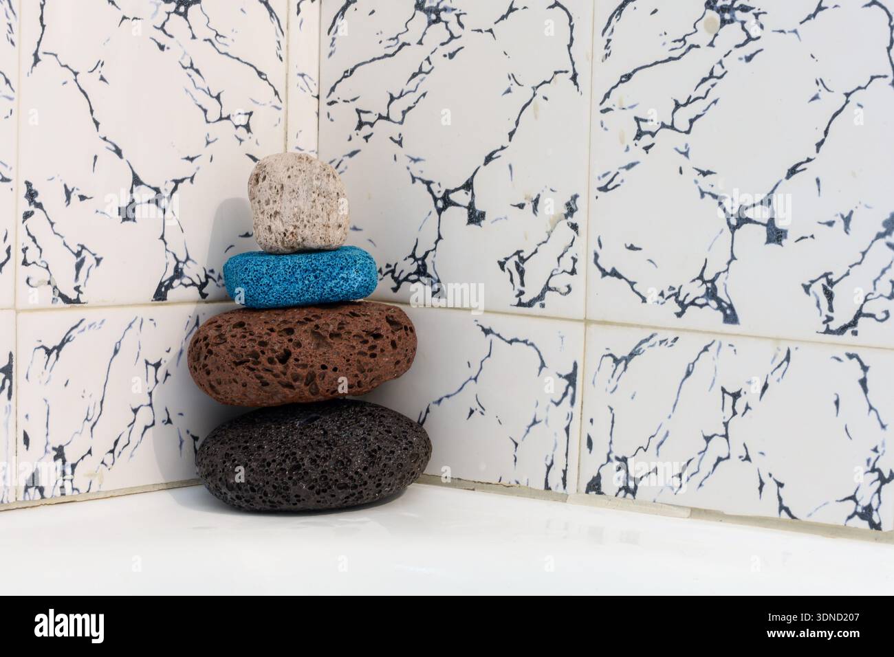 quattro pietre pomice, tre naturali e una sintetica blu brillante impilate in un angolo del bagno su una parete piastrellata con motivi in marmo. Foto Stock