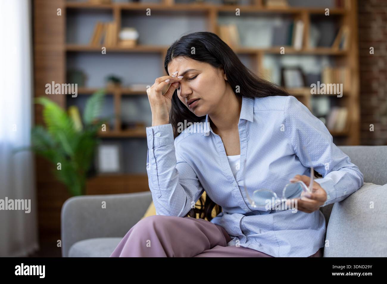 Donna che soffre di grave emicrania e affaticamento degli occhi, che tiene gli occhiali e preme le tempie nel dolore mentre è seduta su un divano, stanca e stressata dal lavoro eccessivo a casa Foto Stock