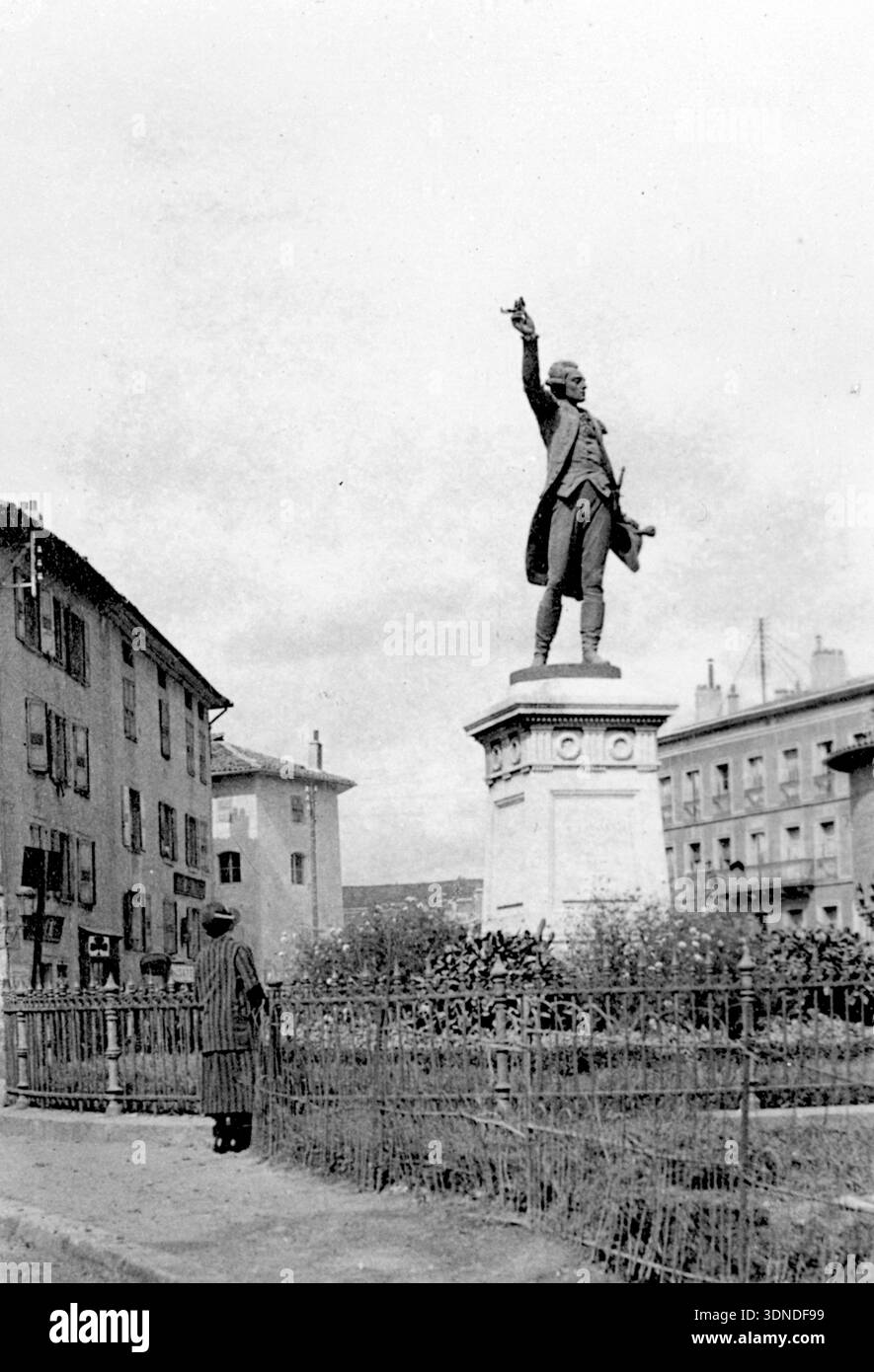 Una statua si erge su un piedistallo decorato all'interno di un giardino recintato al centro di una piazza cittadina. La figura solleva un braccio con un gesto di comando. Gli edifici circostanti, con finestre con persiane, incorniciano lo spazio, mentre un passante solitario con un lungo cappotto si ferma per guardare verso il monumento. Da una collezione di fotografie uniche del XX secolo. Fotografo dilettante, scatti e alcuni più grandi in un singolo album, segnando i viaggi europei di una famiglia. Foto Stock