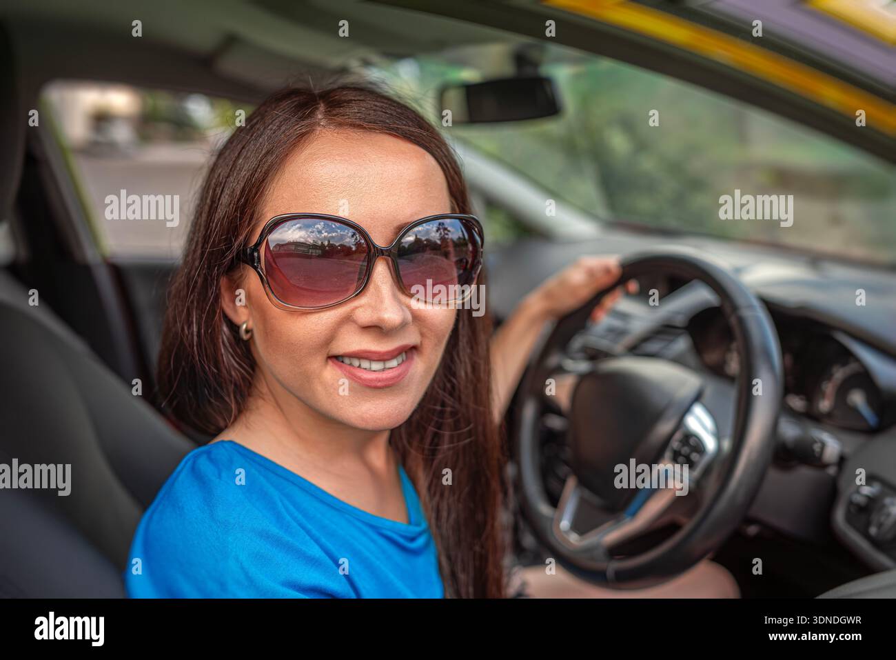 Una giovane donna sorridente con gli occhiali da sole siede al posto di guida di un'auto. Foto Stock