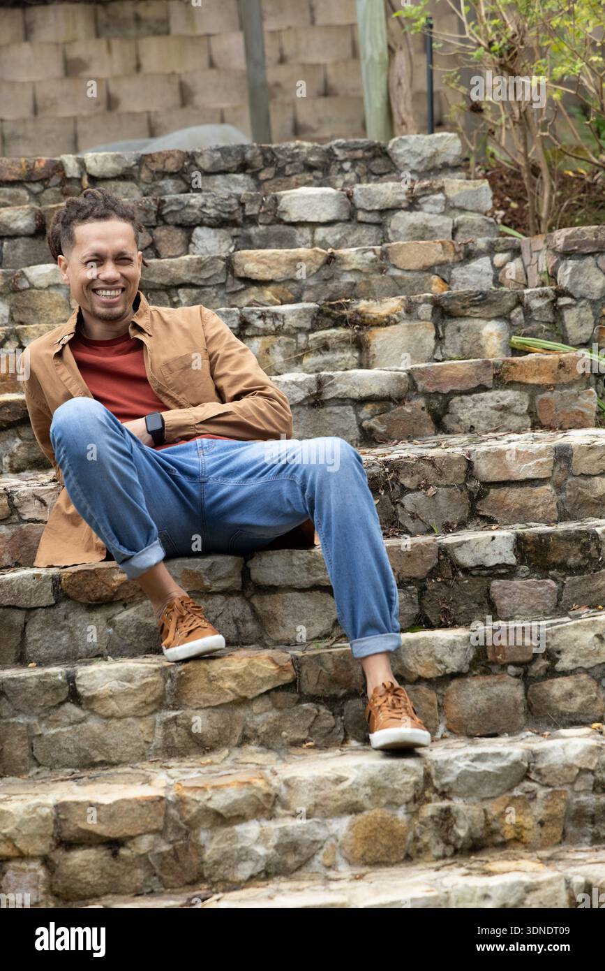 Uomo seduto e appoggiato su gradini di pietra sulla terrazza all'aperto con smartwatch e jeans Foto Stock