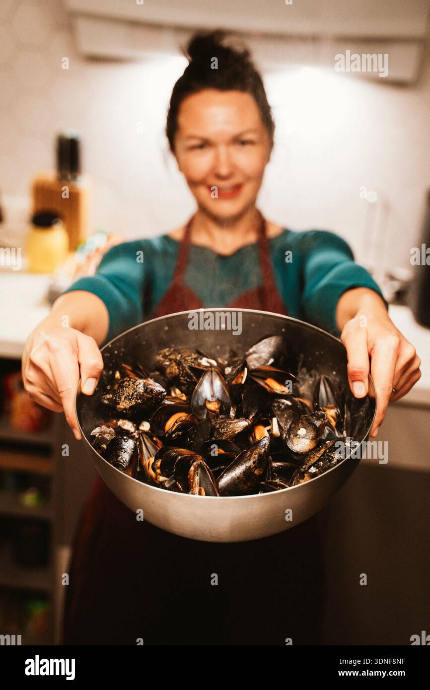 Donna sorridente in cucina che offre una ciotola di cozze al vapore. La scena trasmette un senso di calore e ospitalita', con pesce fresco pronto per essere enj Foto Stock