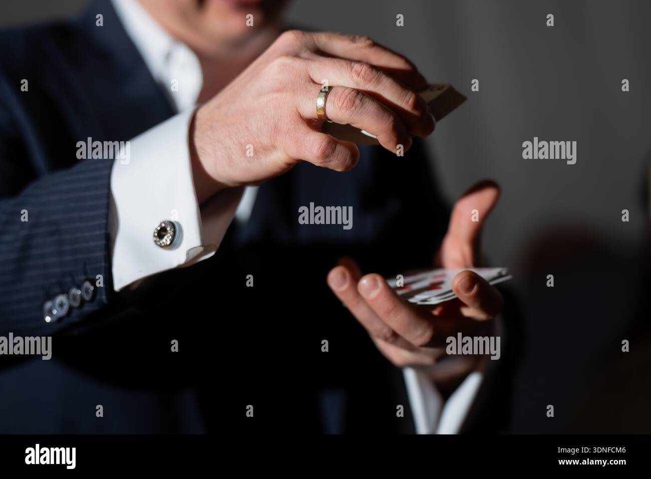 Primo piano del mago delle carte che si occupa e manipola le carte da gioco con le mani, gemelli in tuta e anello d'oro visibili sotto i riflettori Foto Stock