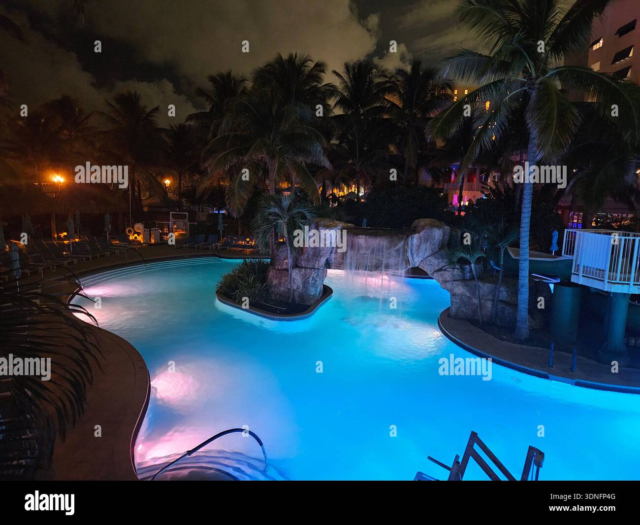 SOLO USO EDITORIALE - MARGARITAVILLE RESORT HOLLYWOOD BEACH FLORIDA: Una vista notturna della piscina in stile laguna del Margaritaville Hollywood Beach Resort, caratterizzato da una prominente cascata di roccia artificiale e da un paesaggio tropicale. La piscina e' illuminata da vibranti luci a LED subacquee blu e rosa, con le ali a piu' piani delle camere ospiti del resort e le silhouette di palme scure sullo sfondo. Questa area ricreativa e' un servizio primario per il resort di destinazione situato sulla Hollywood Beach Broadwalk. Foto Stock