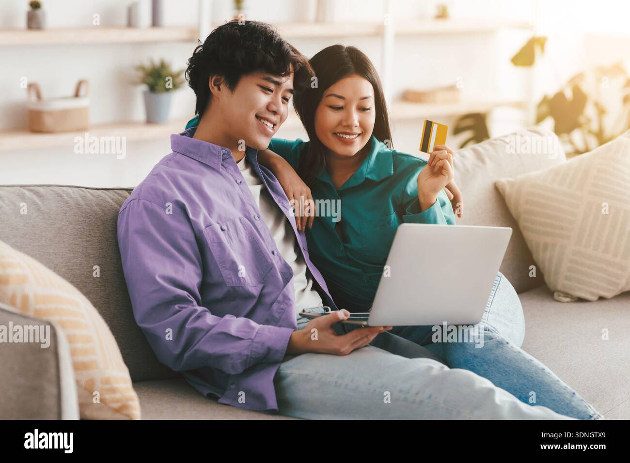 Famiglia asiatica positiva che fa shopping in linea con carta di credito e laptop, seduta sul divano e coccole a casa Foto Stock