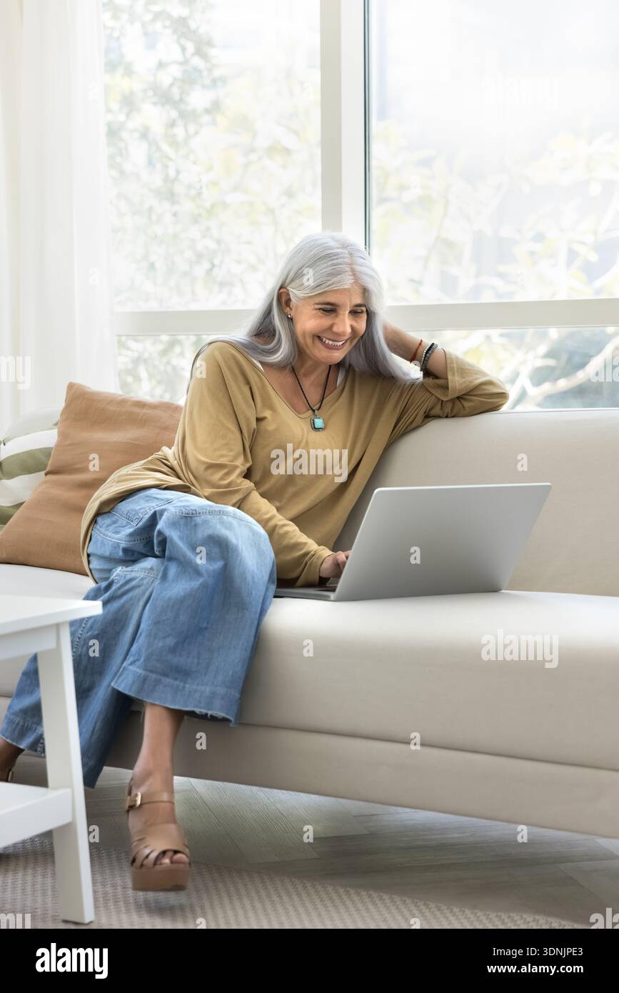 Una donna di mezza età degli anni '50 siede sul divano con un laptop Foto Stock