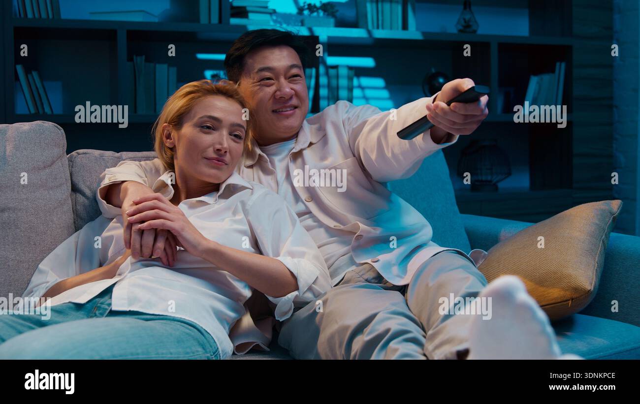 Tranquilla coppia multirazziale che guarda il cinema e la televisione coccolarsi riposando in un comodo soggiorno sul divano la sera a casa di un uomo asiatico eterogeneo Foto Stock