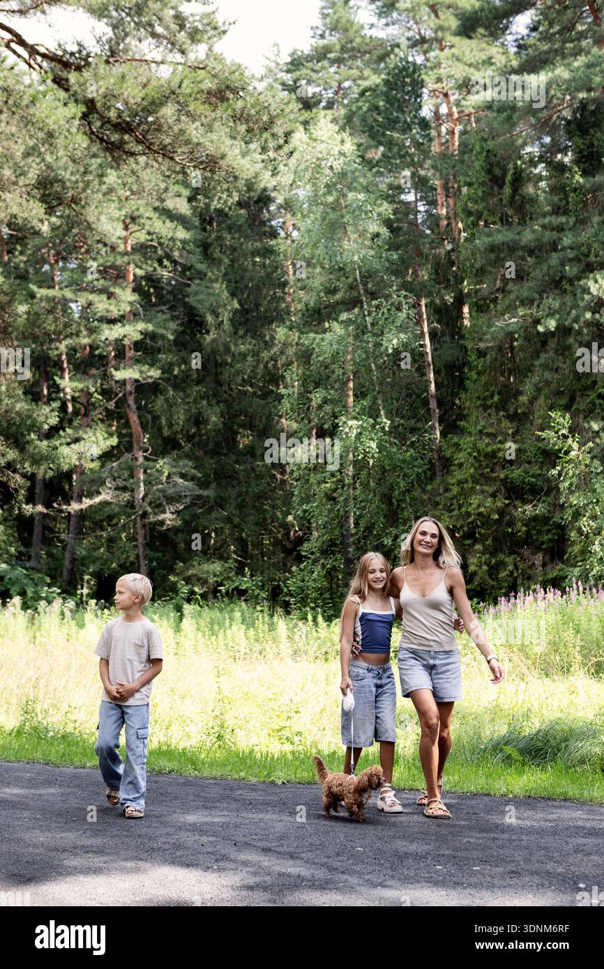 Famiglia felice che cammina insieme a un piccolo cane sul sentiero della foresta. Madre, figlio e figlia sorridenti che godono di un'atmosfera amichevole, insieme, fiducia e.. Foto Stock