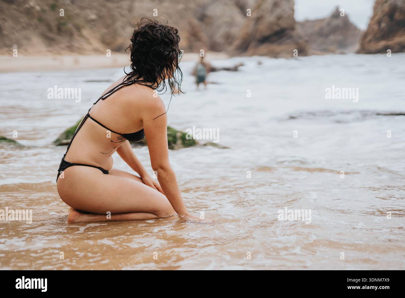 La donna si inginocchia sulla costa in un bikini nero, guardando verso rocce e onde lontane Foto Stock