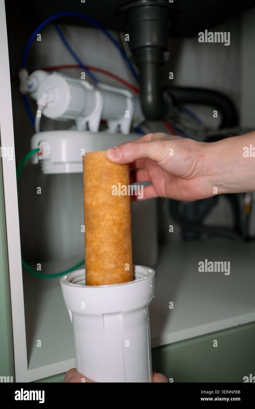 Rimozione manuale di una cartuccia del filtro marrone per sedimenti usata e sporca da un alloggiamento del sistema di osmosi inversa sotto un lavello della cucina, esecuzione di una manutenzione essenziale delle tubazioni e garanzia di acqua potabile pulita Foto Stock