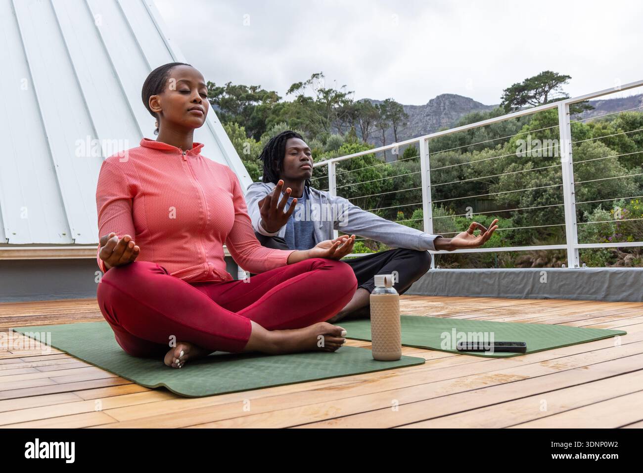 Coppia afroamericana che meditano su tappetini verdi sul tetto, abbigliamento da yoga, bottiglia d'acqua, smartphone Foto Stock
