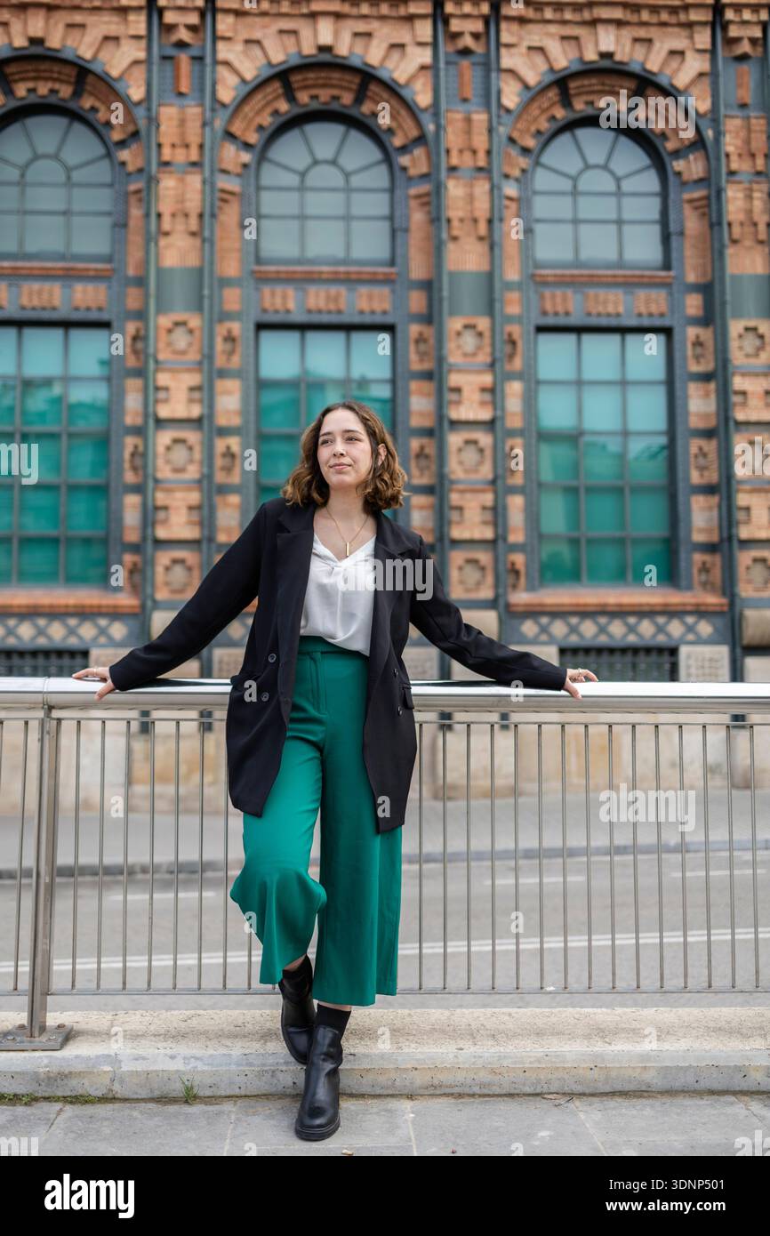Giovane donna appoggiata su una ringhiera di metallo di fronte a un grande edificio in mattoni ornato con finestre ad arco, che indossa una giacca nera e pantaloni verdi, exud Foto Stock