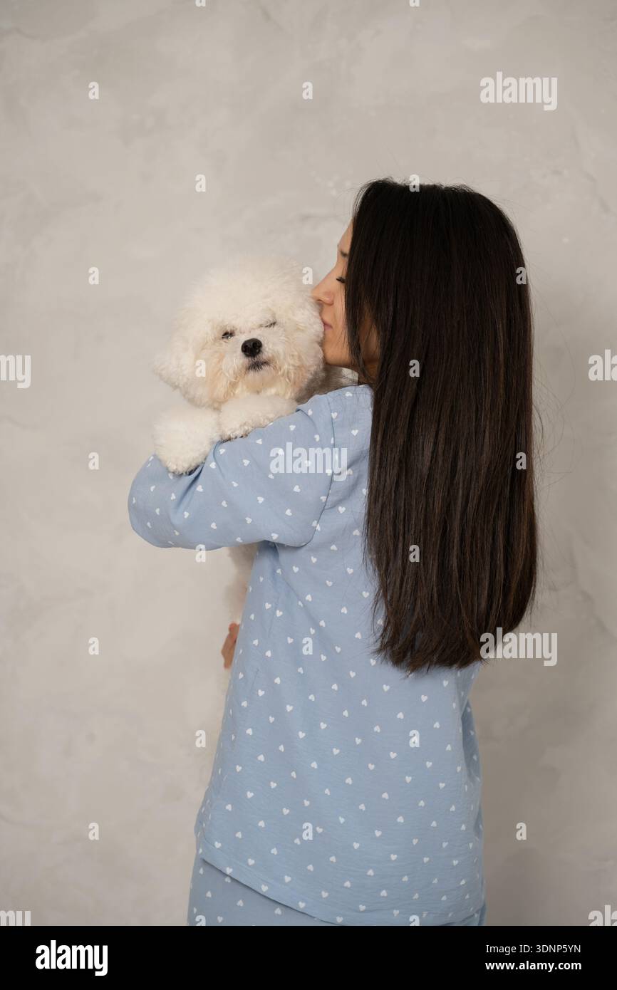 Una donna in un set di pigiama azzurro con motivi a cuore tiene vicino a lei un morbido cane bianco. La scena trasmette calore e comfort in un ambiente domestico. Foto Stock