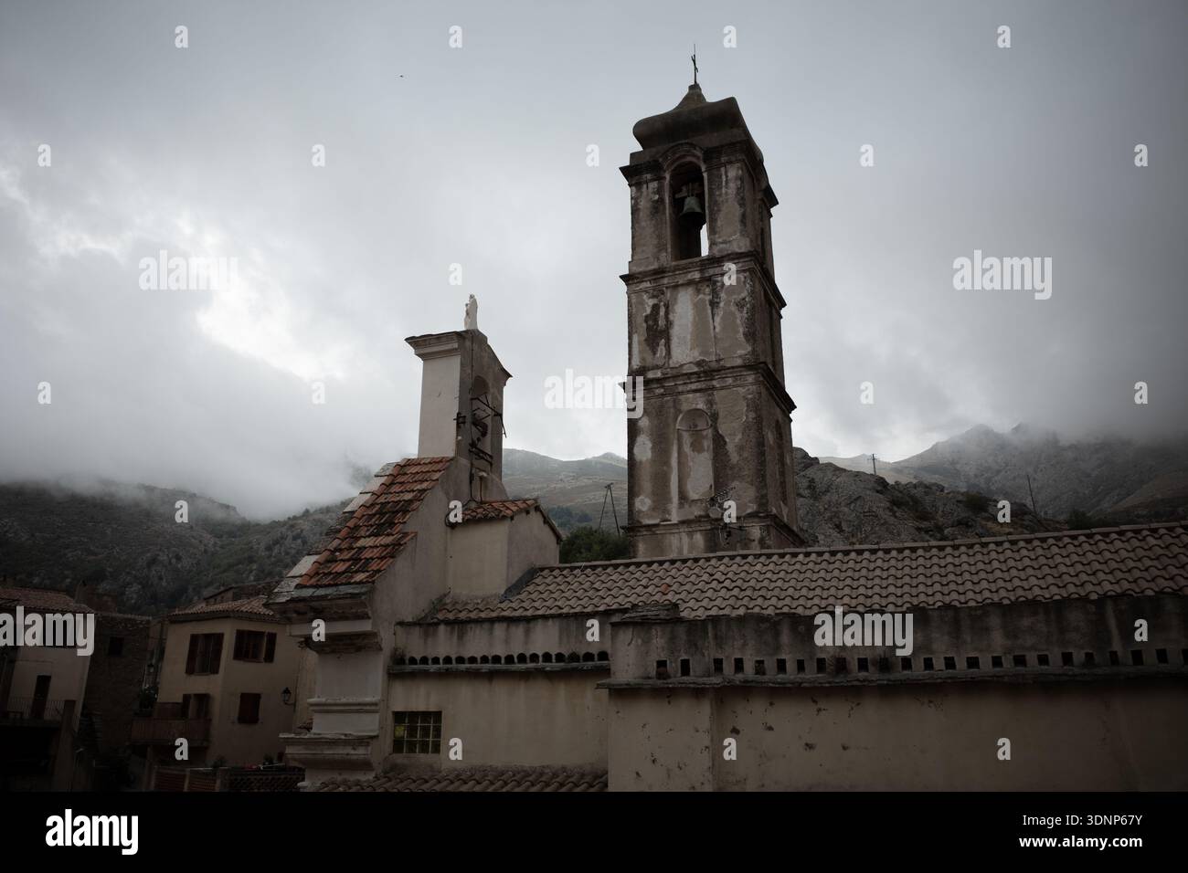 Storica torre della chiesa e tetti piastrellati a Speloncato, Corsica, incorniciati da cime nebbiose sotto il cielo coperto. Foto Stock