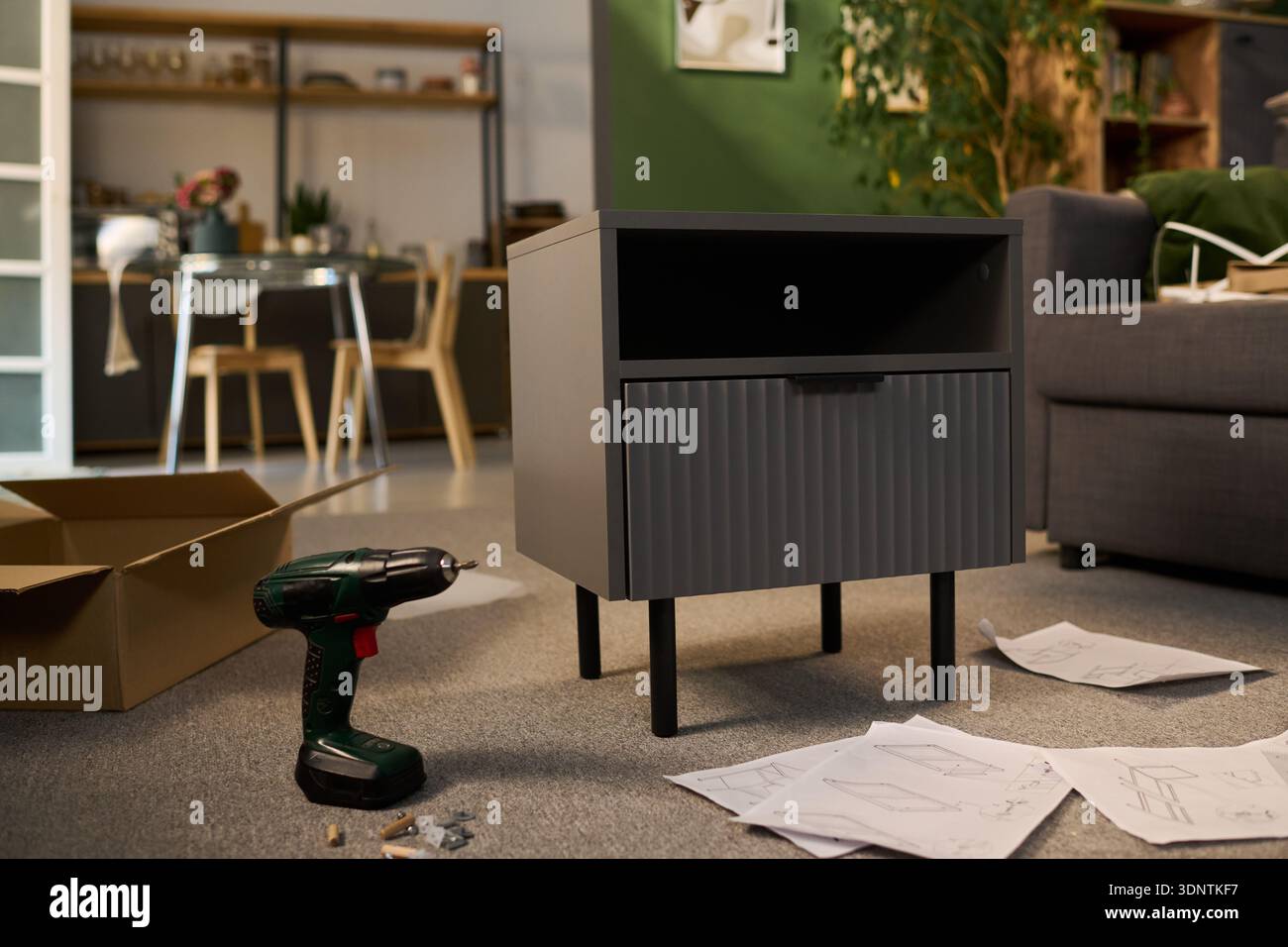 Comodino moderno assemblato in soggiorno con trapano elettrico, viti e fogli di istruzioni sparsi sul pavimento, scatola di cartone visibile sullo sfondo Foto Stock