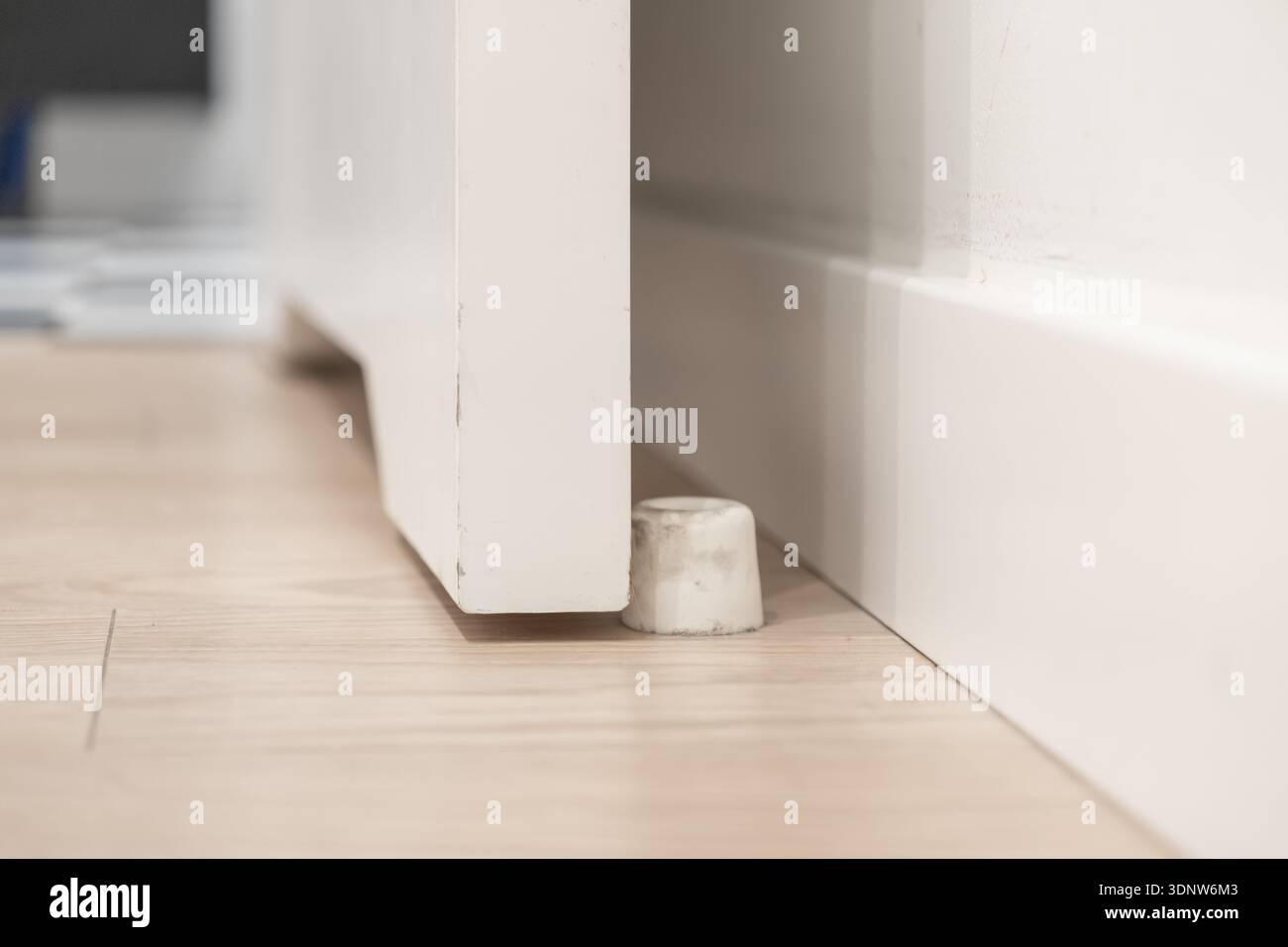 Vista ravvicinata di un piccolo fermaporta bianco cilindrico collocato su un pavimento di legno chiaro, che mantiene una porta bianca minimalista in modo sicuro e socchiusa in un'area residenziale Foto Stock