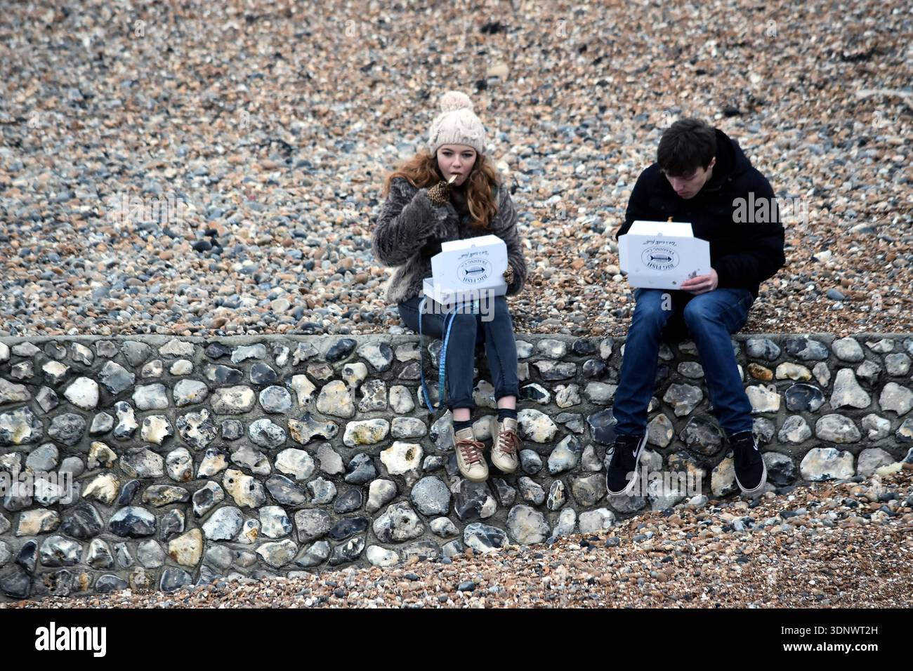 Brighton, Regno Unito - 13 gennaio 2018: Due amici mangiano fish and chips da asporto mentre sono seduti su una parete di ciottoli durante una fredda giornata invernale Foto Stock