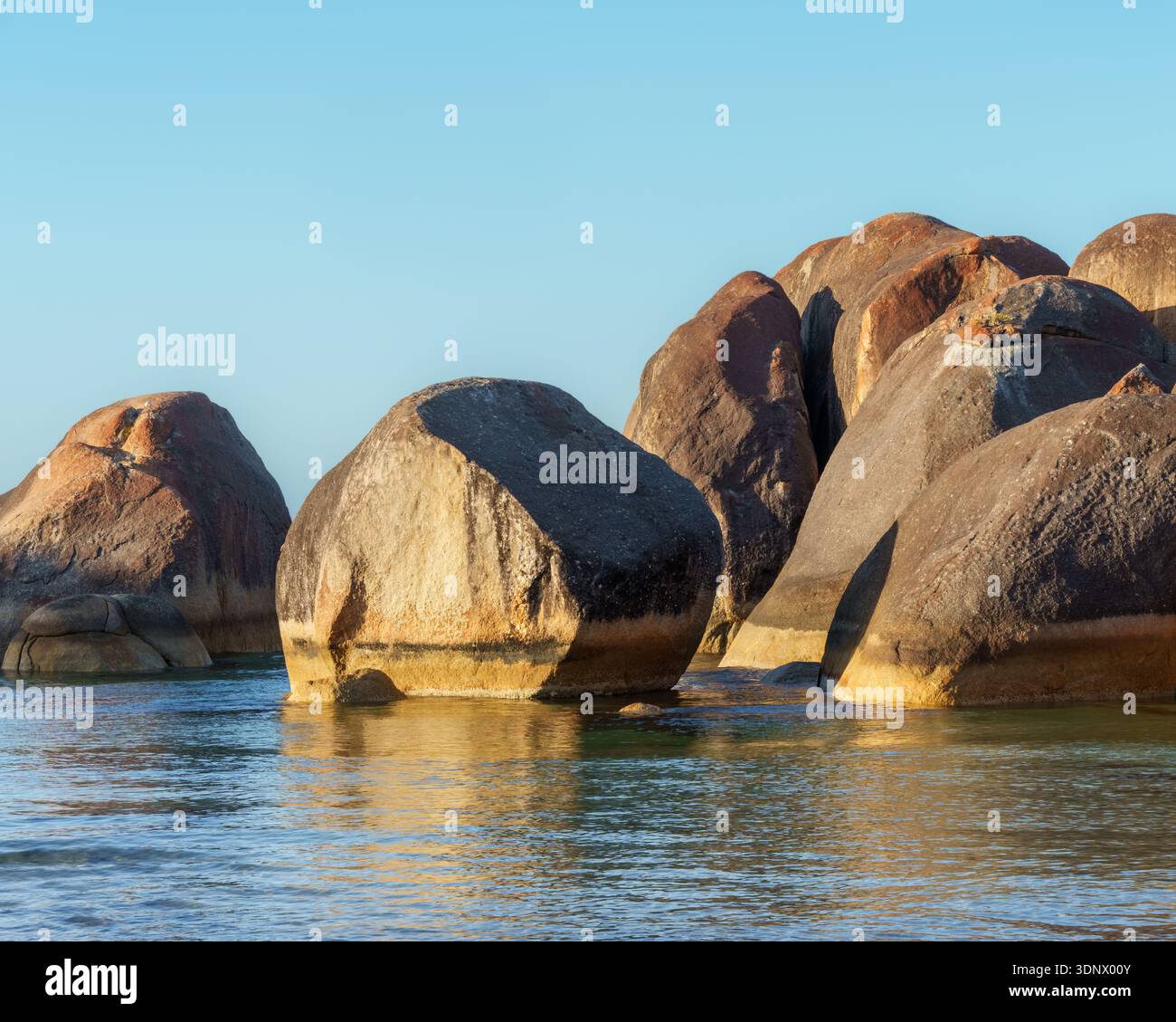 I massi di granito arrotondati si innalzano dall'acqua costiera poco profonda sotto una luce chiara e calda. Foto Stock