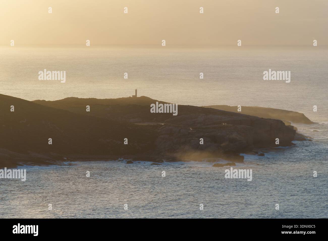 La luce soffusa dell'alba illumina le scogliere costiere frastagliate con la nebbia marina che si innalza dalle onde sottostanti, mentre un piccolo faro si erge su un promontorio lontano. Foto Stock