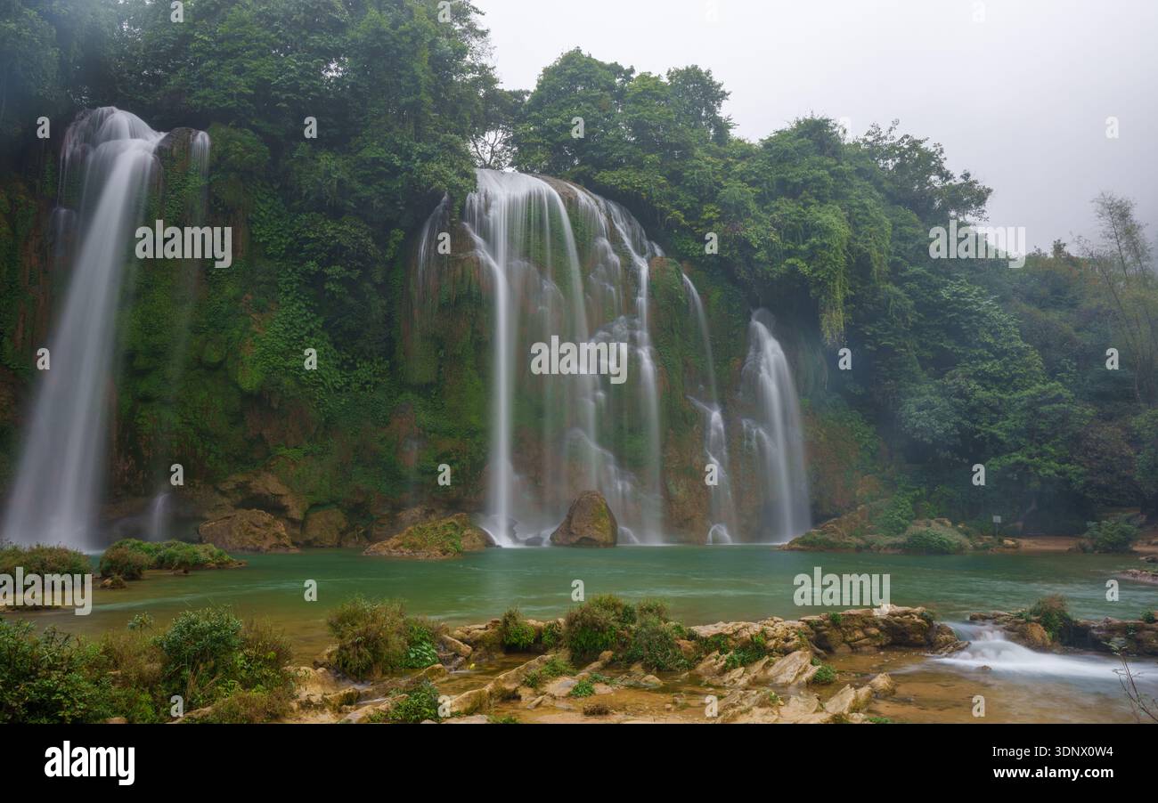 Un'ampia cascata scorre su rocce coperte di muschio in una tranquilla piscina circondata da una fitta vegetazione tropicale all'interno del complesso delle cascate Ban Gioc. Foto Stock