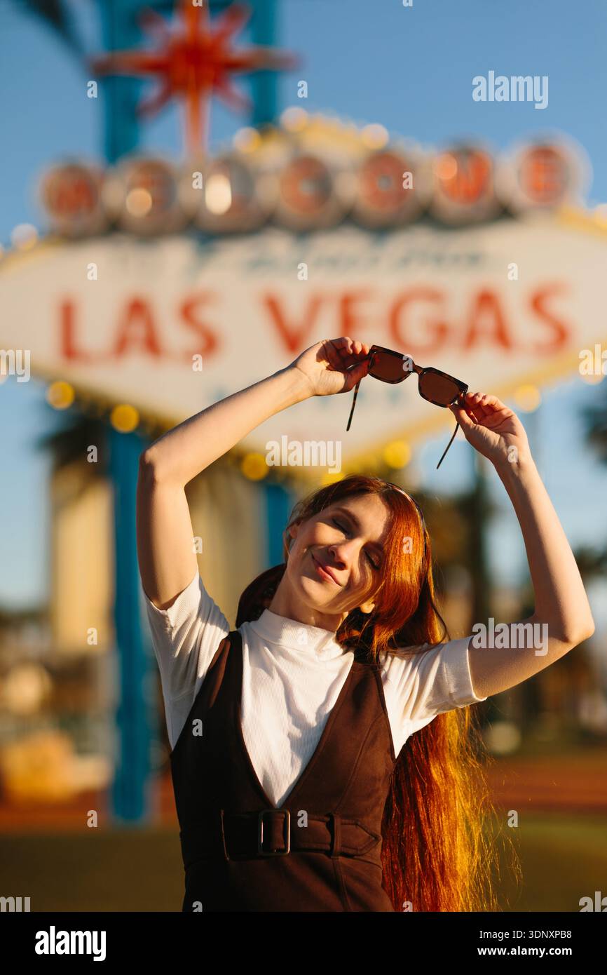 Una giovane donna gioiosa si erge davanti all'iconico cartello di Las Vegas, crogiolandosi alla luce del sole. La sua posa rilassata e il suo sorriso riflettono l'emozione di una visita Foto Stock