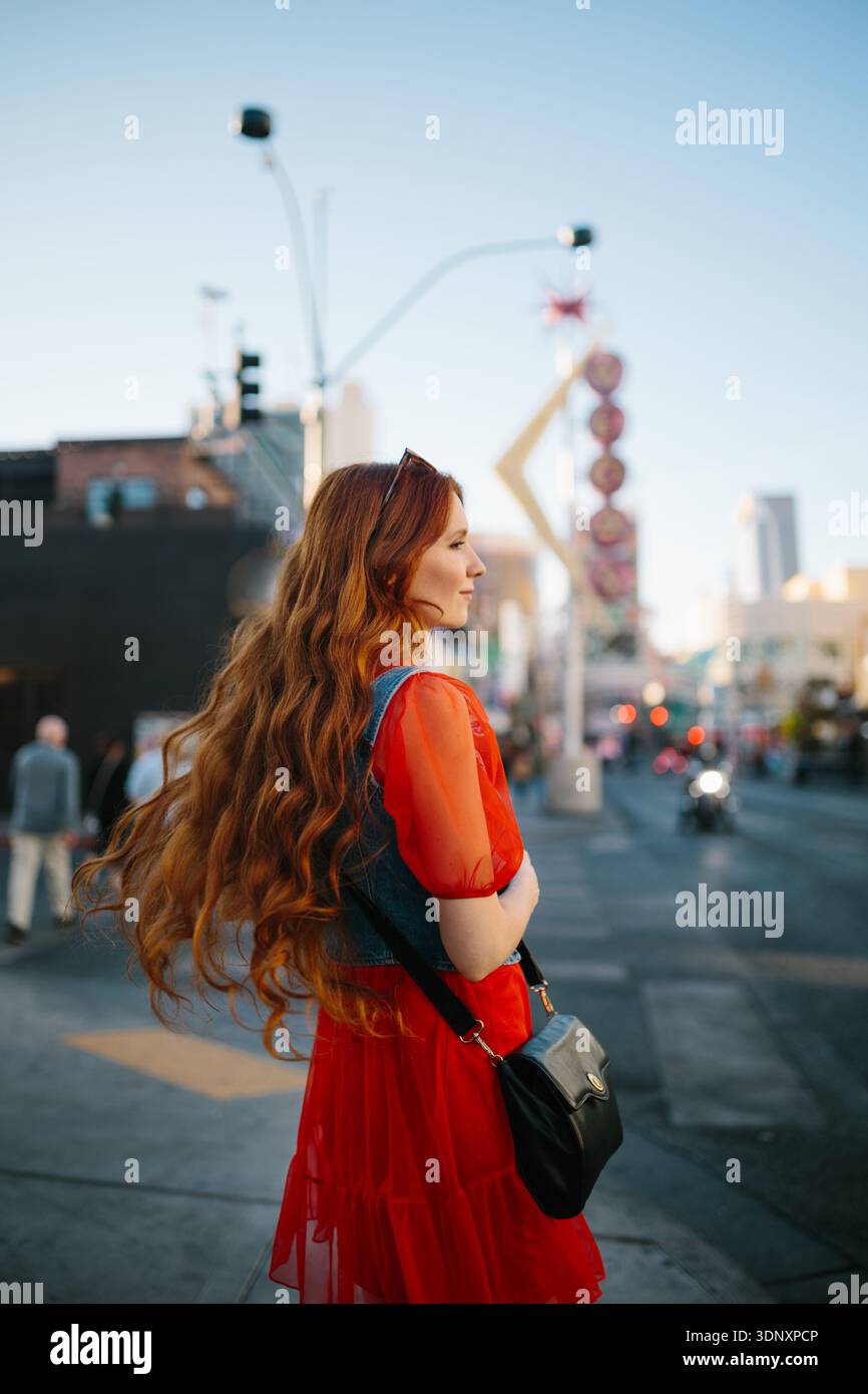 Una giovane donna con i capelli lunghi e un vestito rosso si erge con fiducia in una vivace strada di Las Vegas. Le insegne al neon e il paesaggio urbano creano uno sfondo vivace Foto Stock