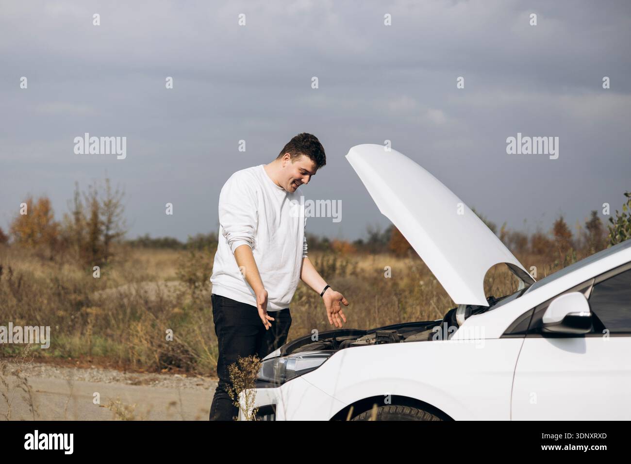 Un giovane uomo si trova di fronte alla sua auto elettrica bianca con il cofano aperto e sembra perplesso. Il veicolo è parcheggiato su una strada di campagna sotto una nuvola Foto Stock