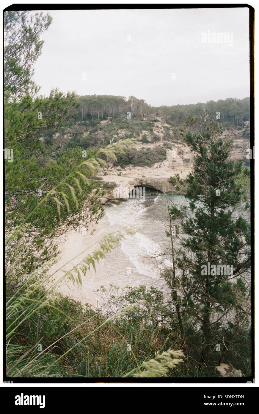 Tranquilla vista costiera di Minorca con scogliere aspre, una baia nascosta e fogliame verde. Le onde del Mar Mediterraneo si infrangono dolcemente sulla spiaggia sabbiosa, e. Foto Stock