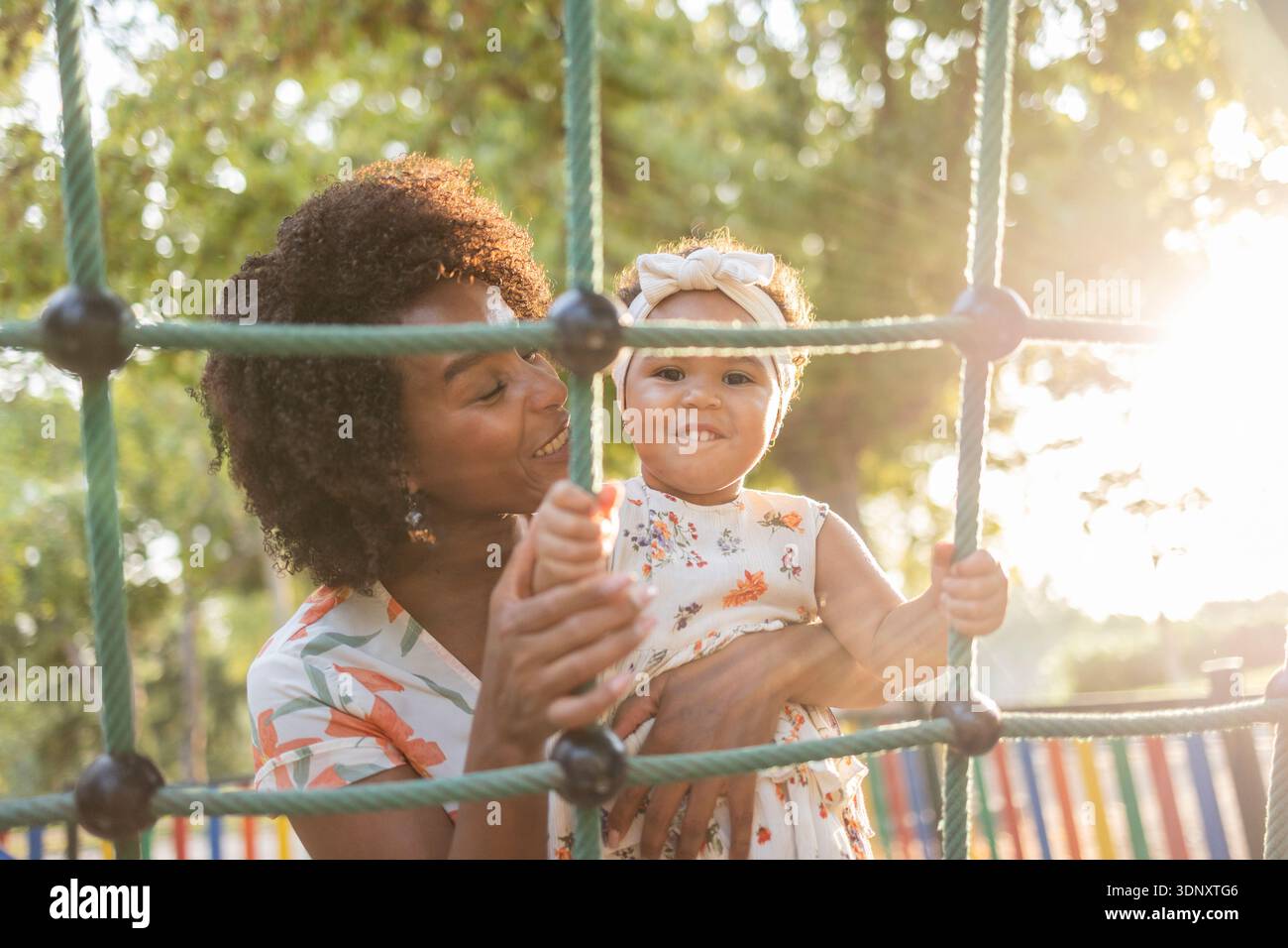 Una madre gioiosa che gioca con il suo bambino in una giornata di sole in un parco giochi, circondato da una vegetazione lussureggiante. Il bambino si aggrappa alla rete per arrampicarsi con un ruscello Foto Stock