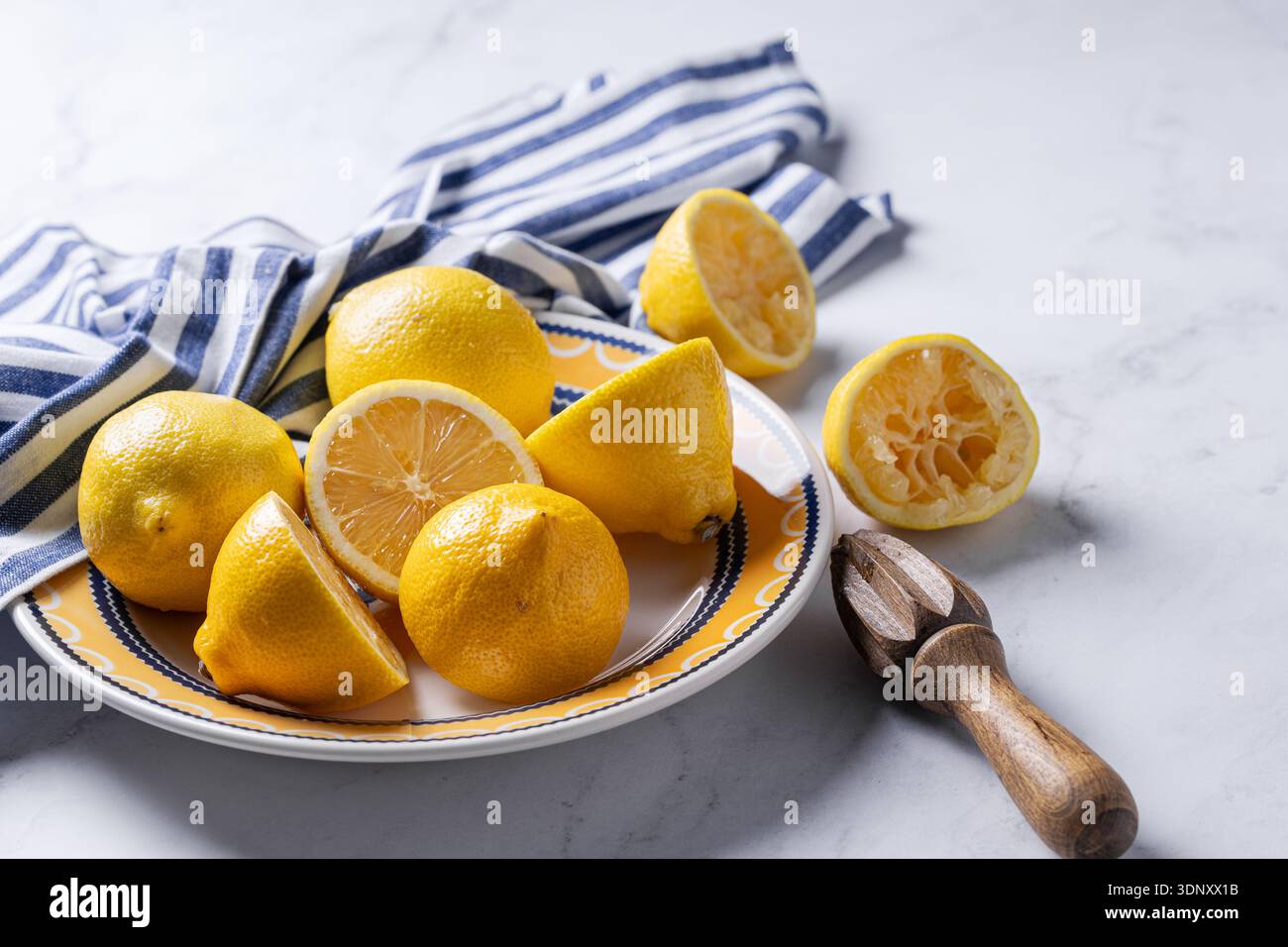 Limoni interi e a fette disposti su un piatto a motivi arancioni con un panno a strisce blu, appoggiato su un piano di marmo accanto a un alesatore di agrumi in legno. Foto Stock