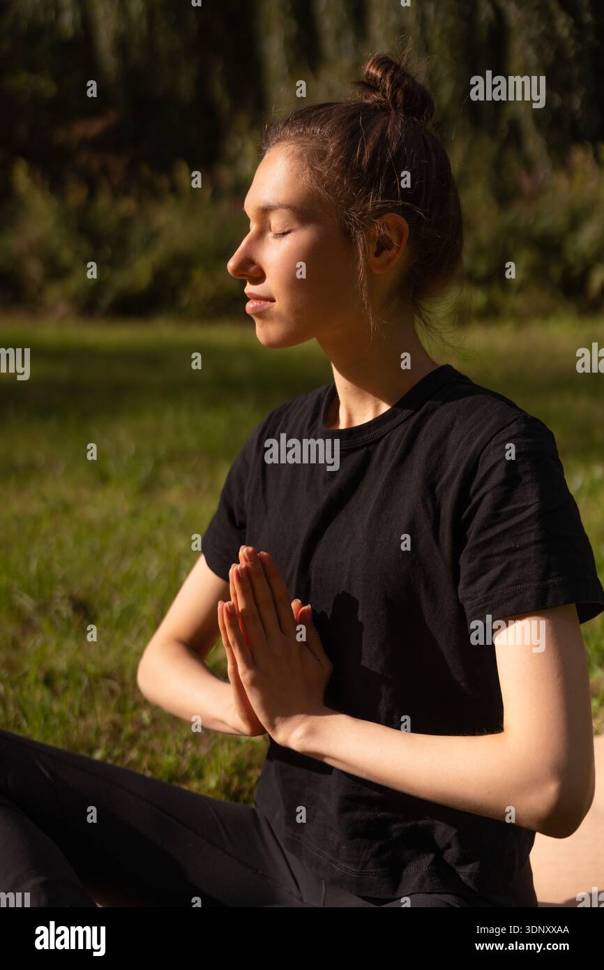 Una giovane donna medita in una posa yoga all'aperto, crogiolandosi alla luce del sole. La sua espressione serena e gli occhi chiusi trasudano tranquillità, mescolandosi con il lussureggiante Foto Stock