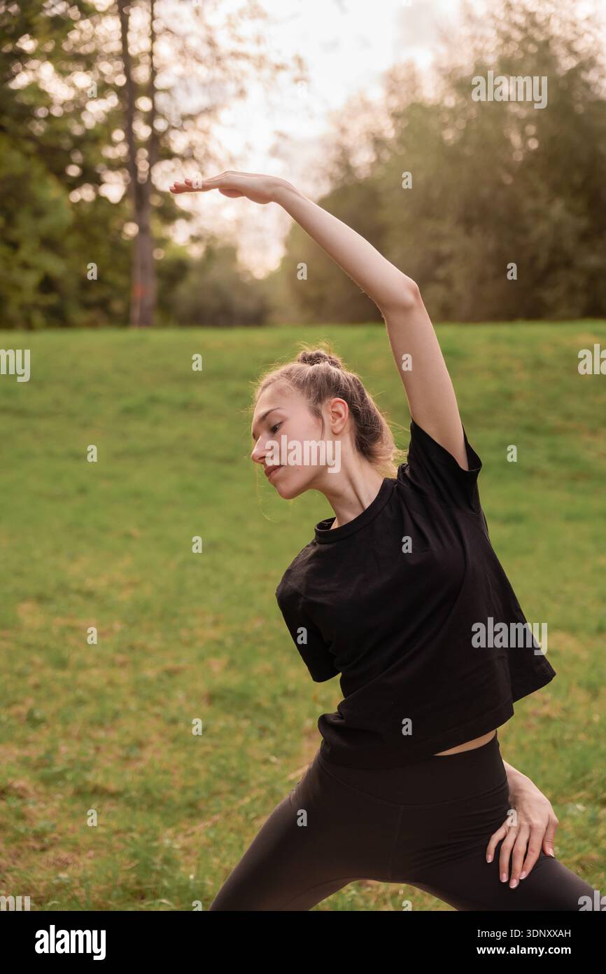 Una giovane donna pratica yoga in un tranquillo ambiente all'aperto, circondato da lussureggianti prati verdi e alberi. Tiene con grazia una posa yoga, incarnando tran Foto Stock