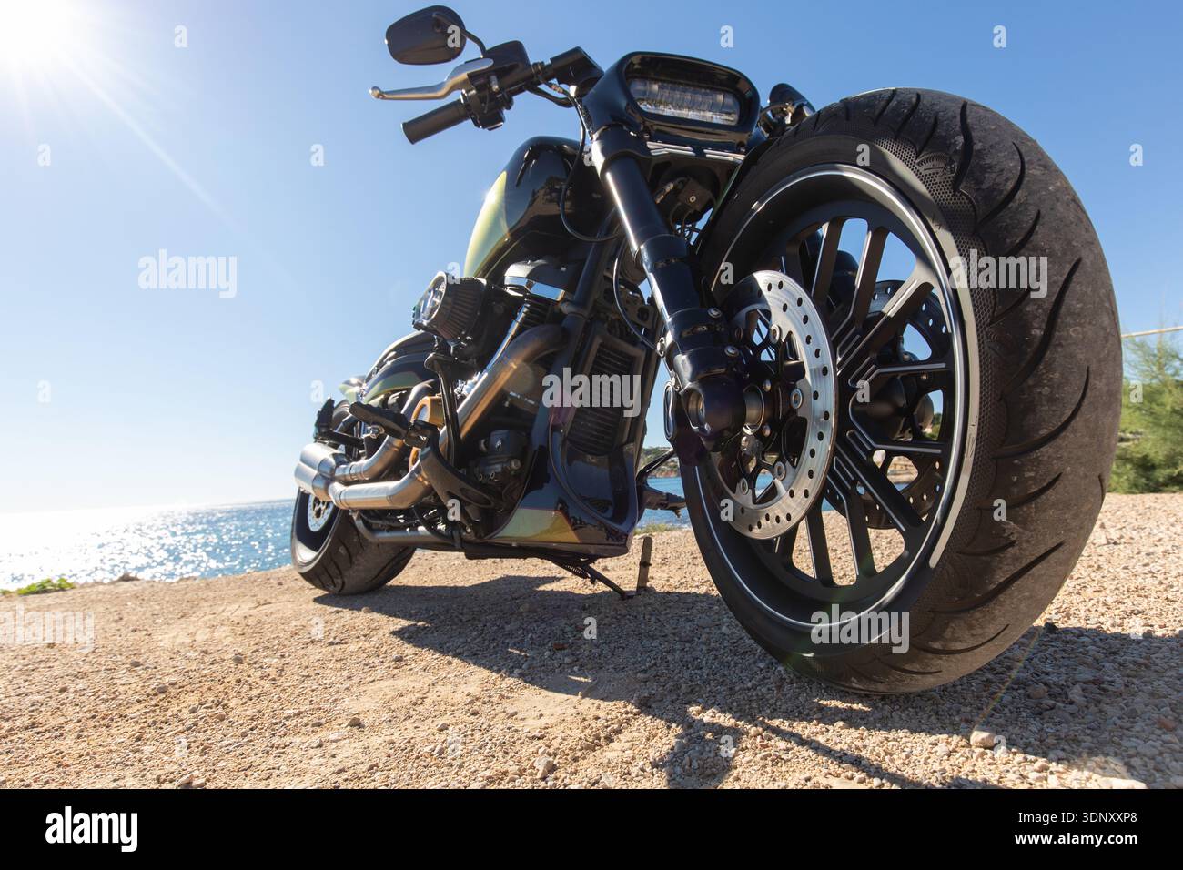 Una motocicletta personalizzata è parcheggiata su un sentiero ghiaioso vicino al mare, con il sole che splende brillantemente. Il design unico e la potente tecnologia della moto evocano un'esperienza unica Foto Stock