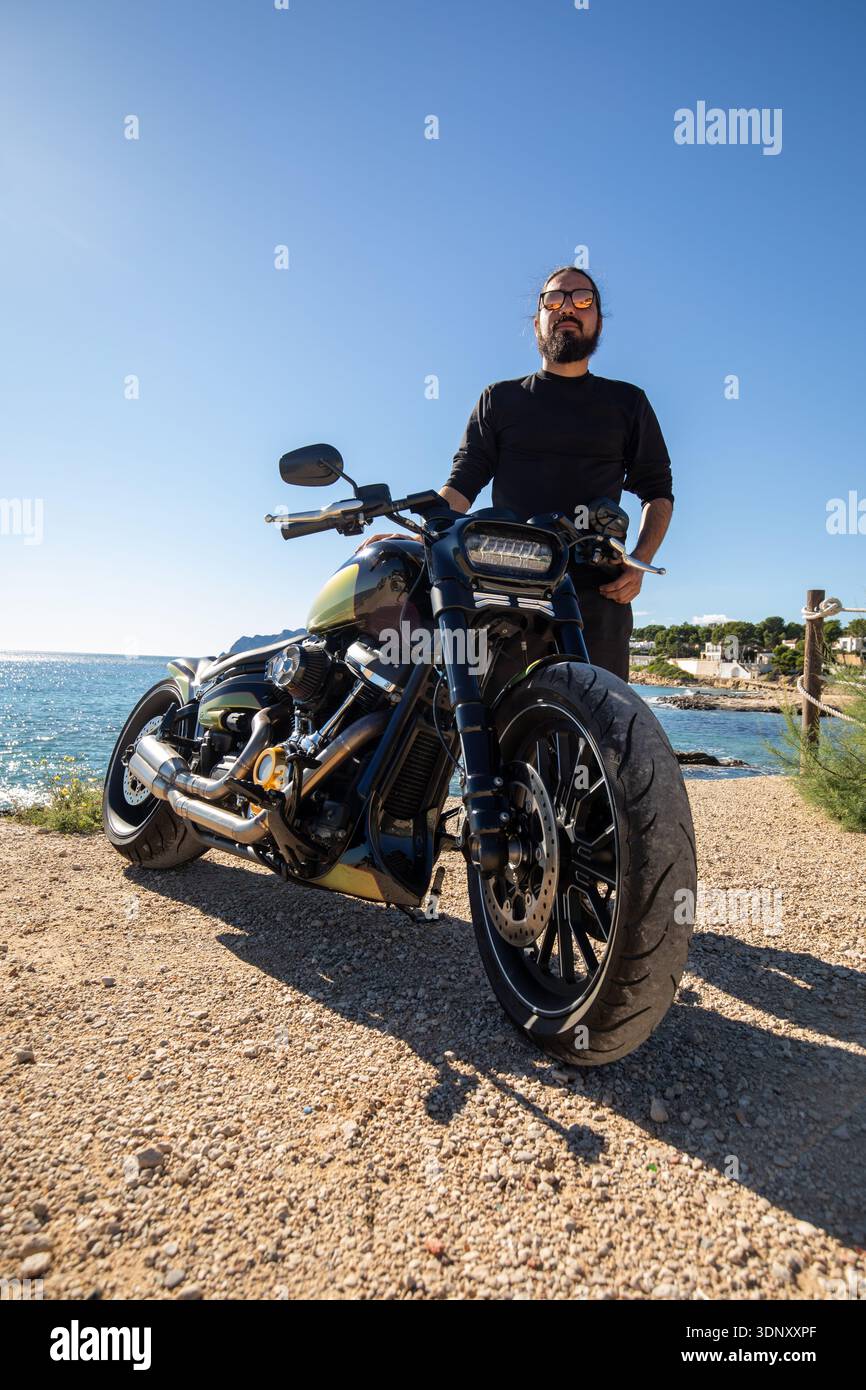 Un'elegante motocicletta personalizzata parcheggiata su una soleggiata strada sulla spiaggia, con un avventuroso motociclista accanto, pronta ad esplorare il paesaggio costiero sotto Foto Stock