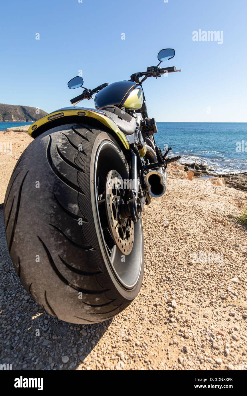 Una motocicletta personalizzata parcheggiata lungo un percorso costiero panoramico, con la sua grande ruota posteriore in contrasto con lo sfondo blu dell'oceano, invitando un avvincente annuncio Foto Stock