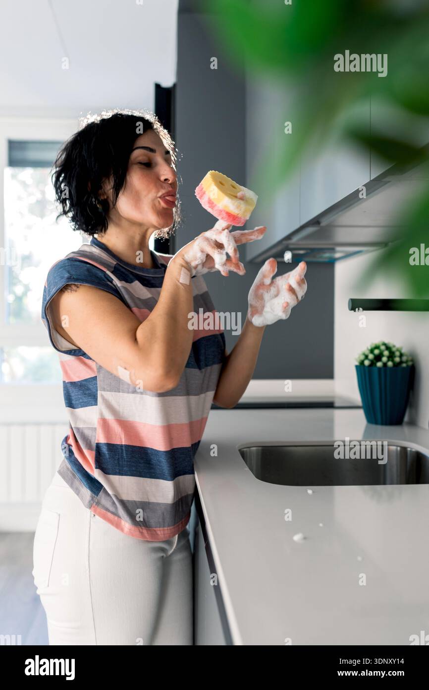 Una donna con un abbigliamento casual bilancia giocosamente una spugna colorata al dito in una cucina moderna, con il viso che crea un'espressione divertente vicino a un lavandino. Foto Stock