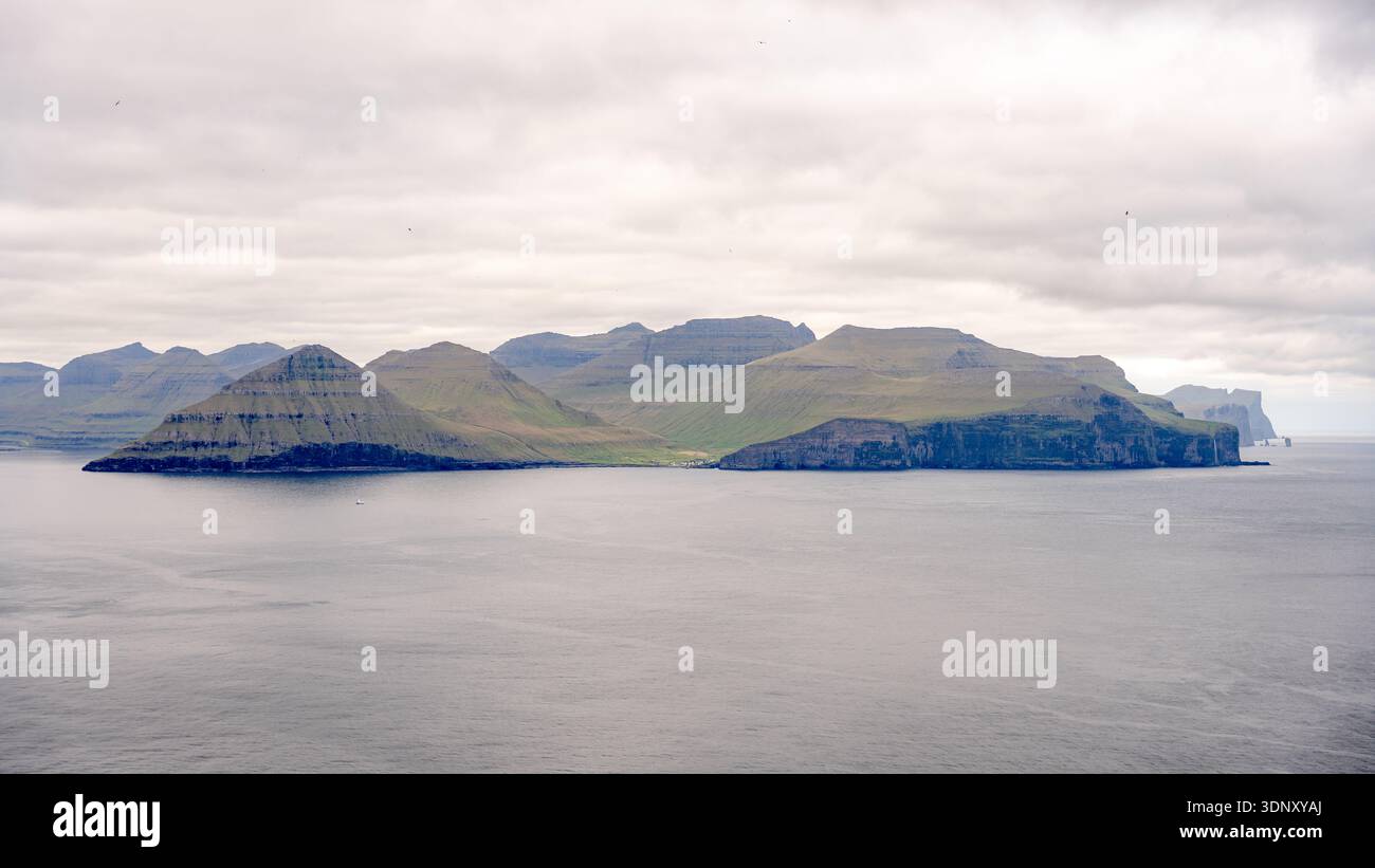 Tranquillo paesaggio di Gjogv, un affascinante villaggio di Eysturoy, Isole Faroe. Questa vista panoramica cattura le maestose scogliere e il mare tranquillo sotto un cl Foto Stock