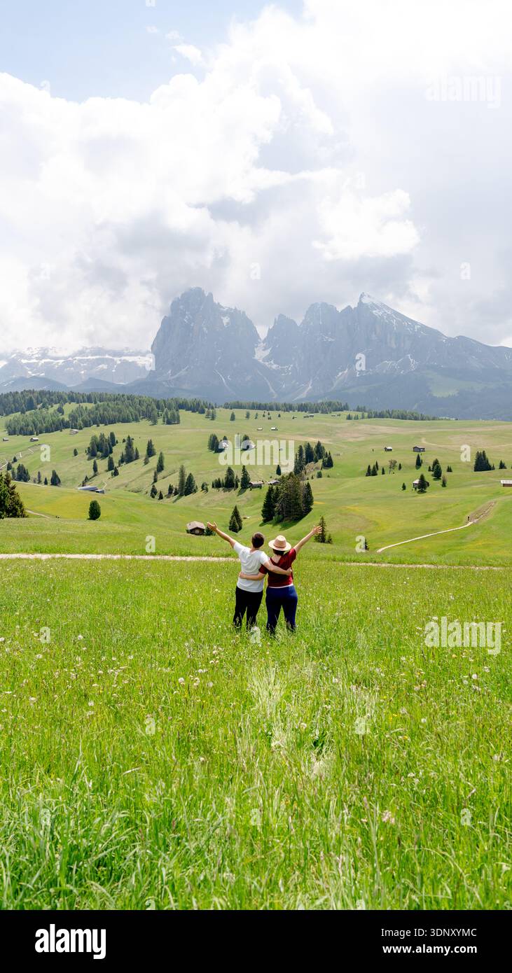 Due persone godono di una vista mozzafiato sui verdi prati lussureggianti e le vette torreggianti dell'Alpe di Siusi, dell'Alpe di Siusi, nelle Dolomiti, in Italia, sotto un dramma Foto Stock