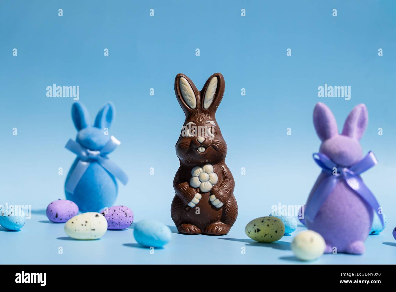 Un coniglietto pasquale al cioccolato si erge orgogliosamente tra coniglietti colorati di feltro blu e viola con nastri. Le uova macchiate dai colori vivaci sono sparse su una p Foto Stock