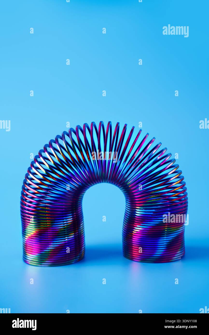 Una colorata molla in metallo dalle tonalità neon forma un arco su uno sfondo blu brillante. Il design astratto mette in risalto l'affascinante gioco di luci e. Foto Stock