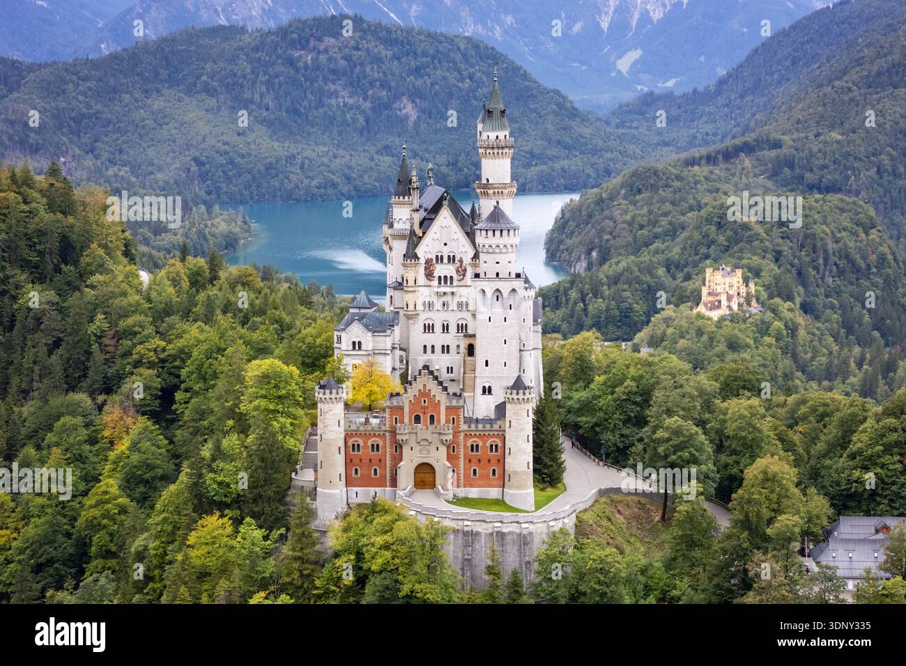 Vista aerea delle maestose torri del castello di Neuschwanstein che si innalzano nel cielo, contrastando con le foreste verdeggianti e il blu sereno dell'Alpse Foto Stock