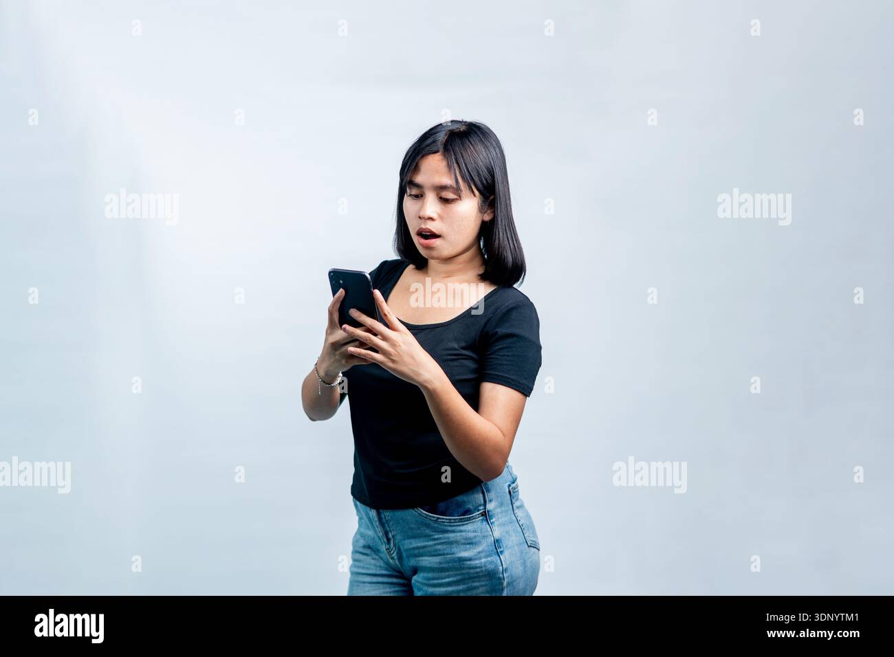 Una giovane donna con una t-shirt nera che guarda il suo telefono con un'espressione scioccata a bocca aperta su uno sfondo bianco pulito. Foto Stock