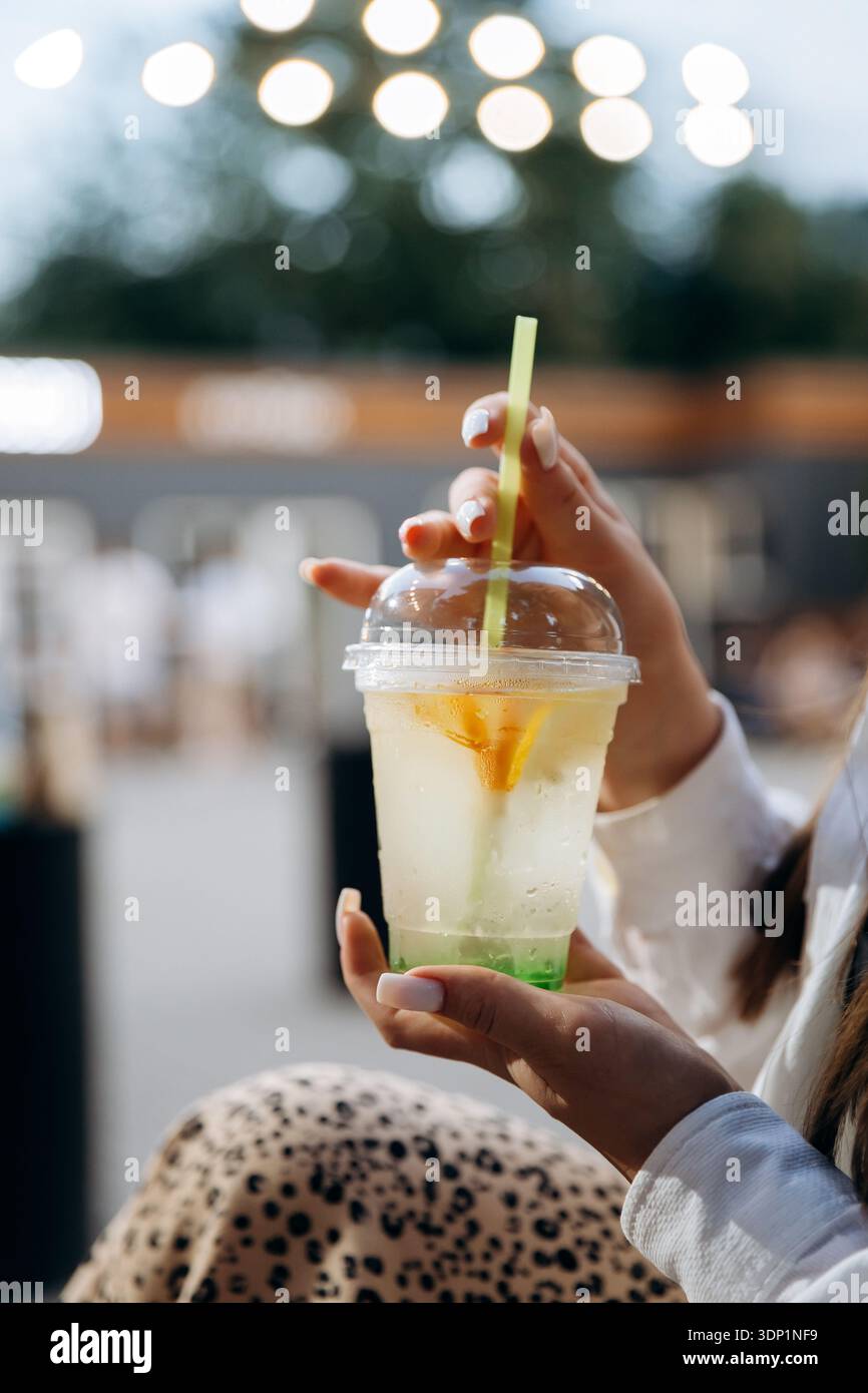 Donna che tiene una bevanda al limone ghiacciata in una tazza di plastica con paglia, caffè bokeh all'aperto Foto Stock