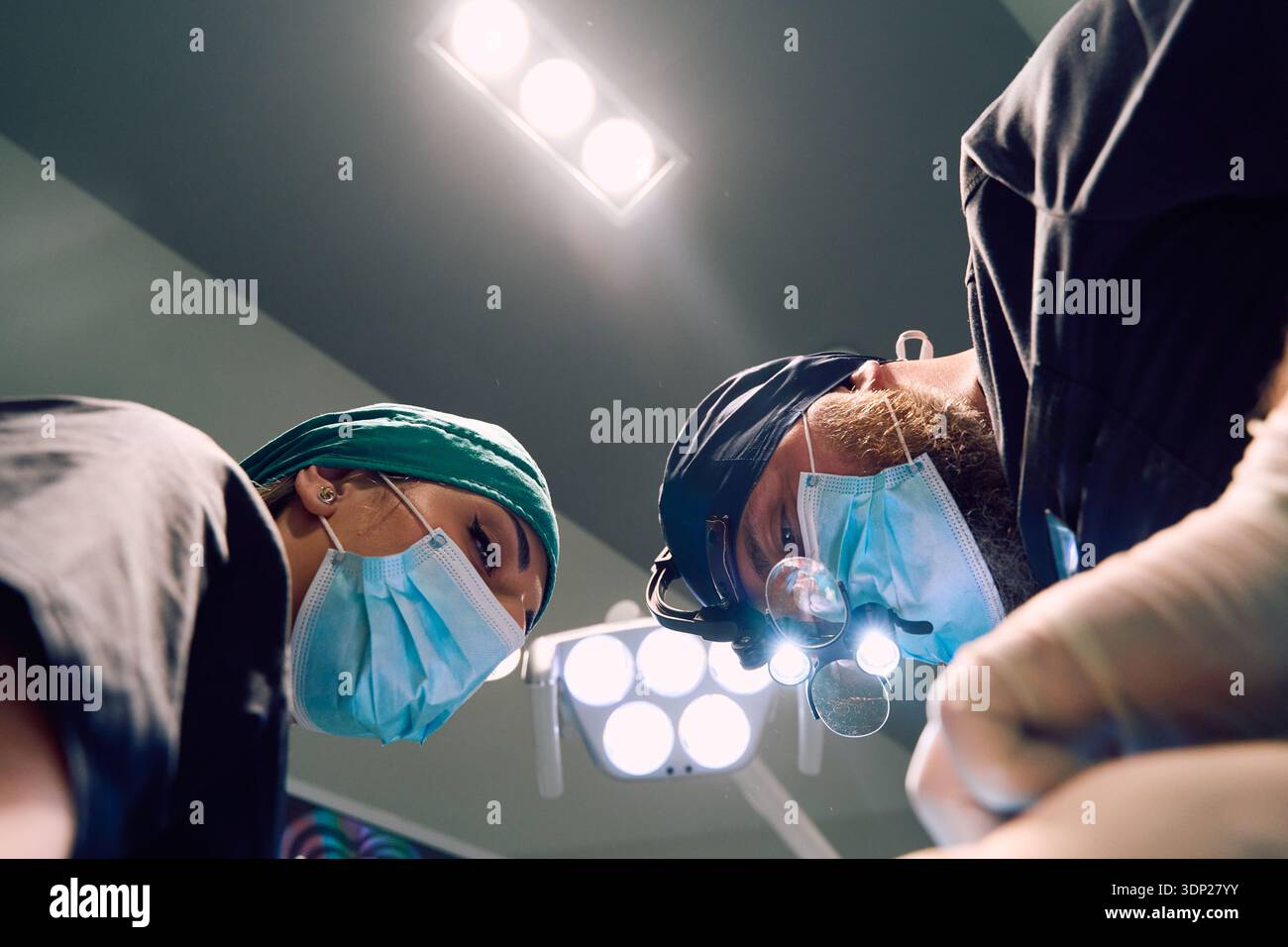 Chirurghi in maschera e cappuccio sotto le luci a soffitto che eseguono interventi chirurgici in sala operatoria focalizzati sul piano clinico Foto Stock