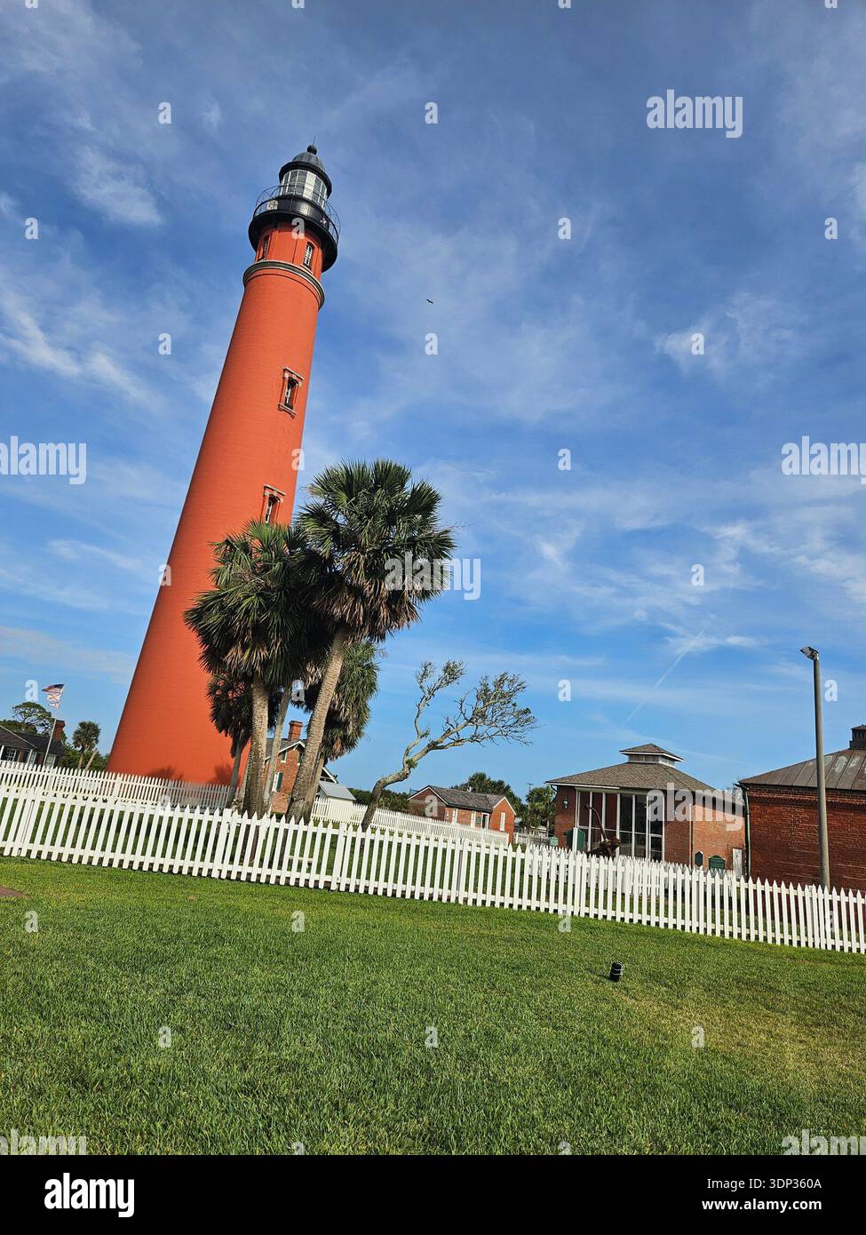 SOLO A USO EDITORIALE - DAYTONA BEACH FLORIDA: ​A vista dall'angolo basso dello storico faro di Ponce de Leon Inlet in Florida sotto un cielo blu con nuvole mostruose. Il faro di mattoni rossi, uno dei più alti degli Stati Uniti, è incorniciato da palme e da una recinzione bianca per picchetti nel parco del museo. - Immagine stock catturata con smartphone