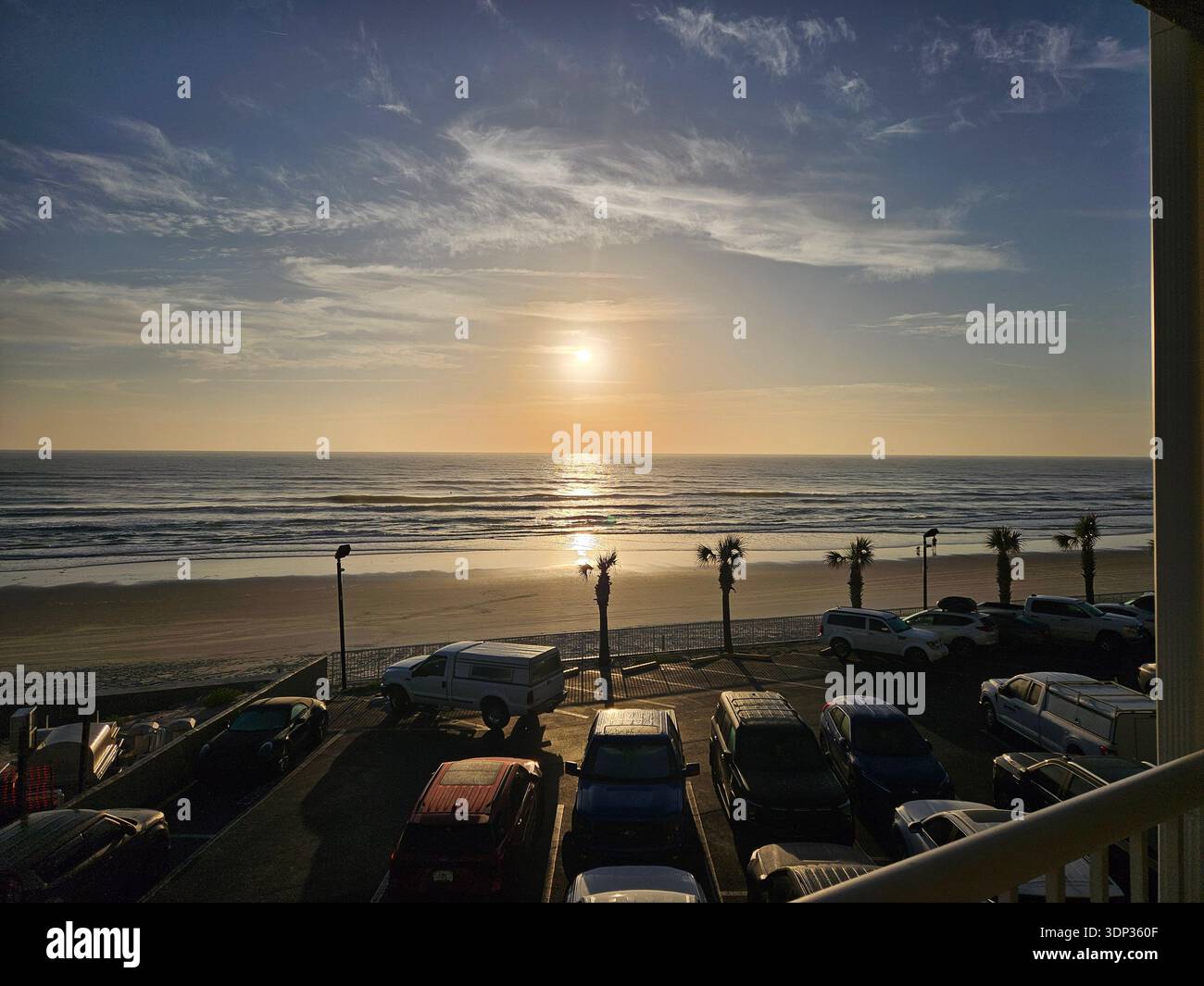 SOLO USO EDITORIALE - DAYTONA BEACH FLORIDA: Una serena vista del tramonto sull'Oceano Atlantico da un balcone fronte spiaggia in Florida. La foto cattura il sole che si riflette sulle onde dell'oceano, una spiaggia sabbiosa costeggiata da palme e un parcheggio costiero in primo piano durante l'ora d'oro. - Immagine stock catturata con smartphone