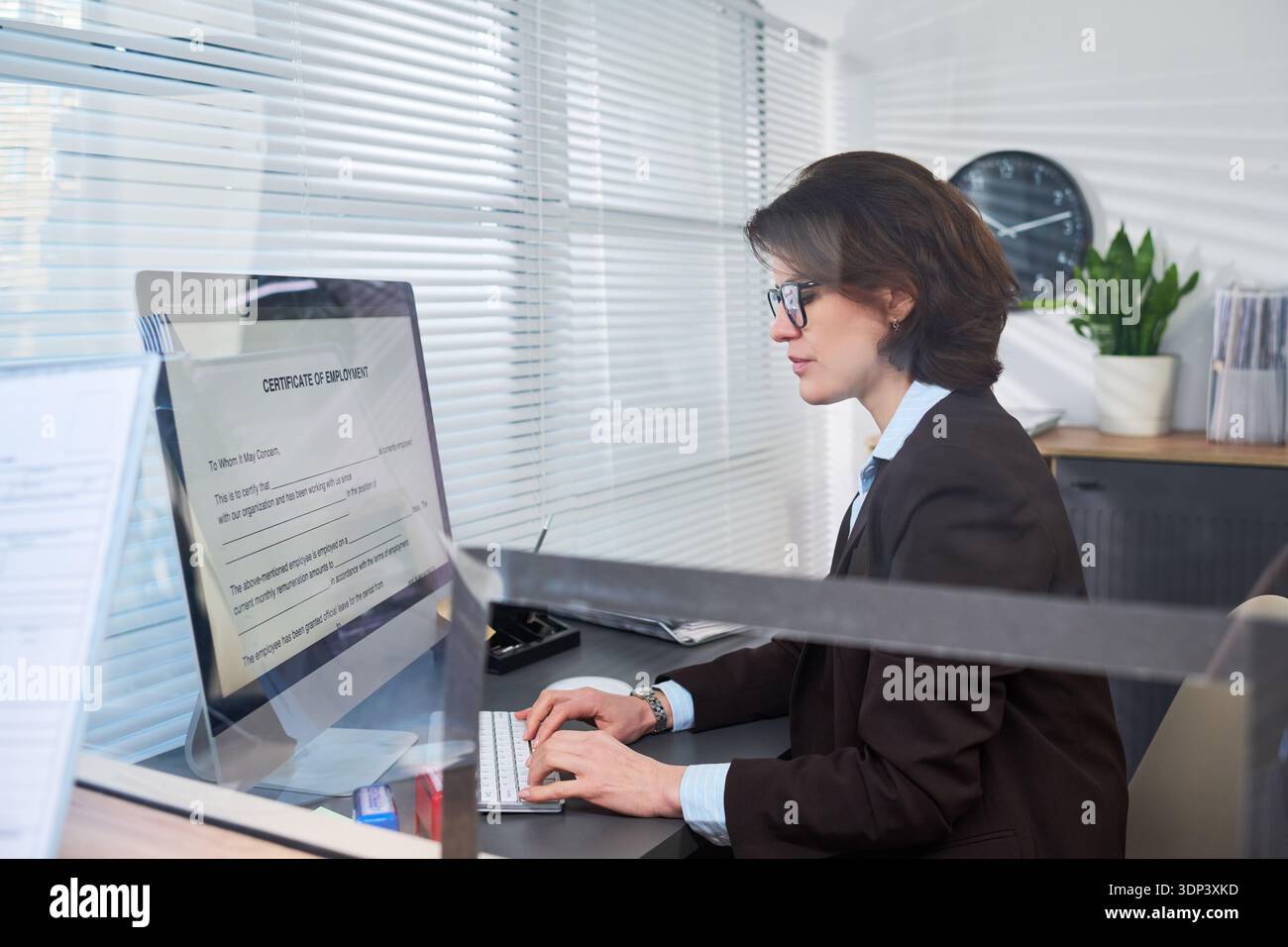 Donna caucasica che lavora alla scrivania nel centro visti digitando sulla tastiera del computer, leggendo il certificato di lavoro visualizzato sul monitor, forniture per ufficio in background Foto Stock
