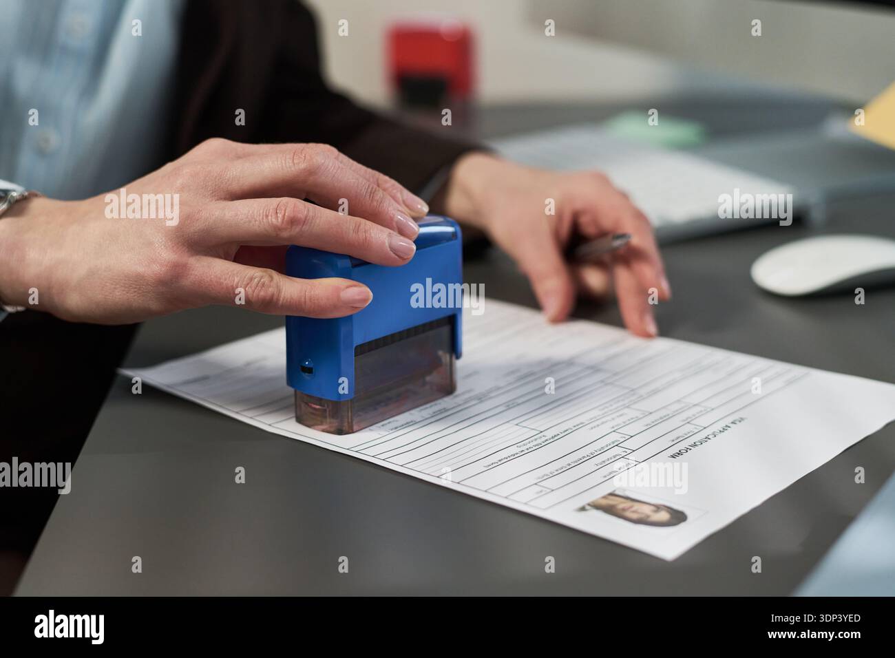 Donna di mezza età sconosciuta che timbra il documento di domanda di visto al centro visti, tiene la penna in mano, ravvicinata delle mani che elabora i documenti sulla scrivania con una foto di identificazione visibile Foto Stock